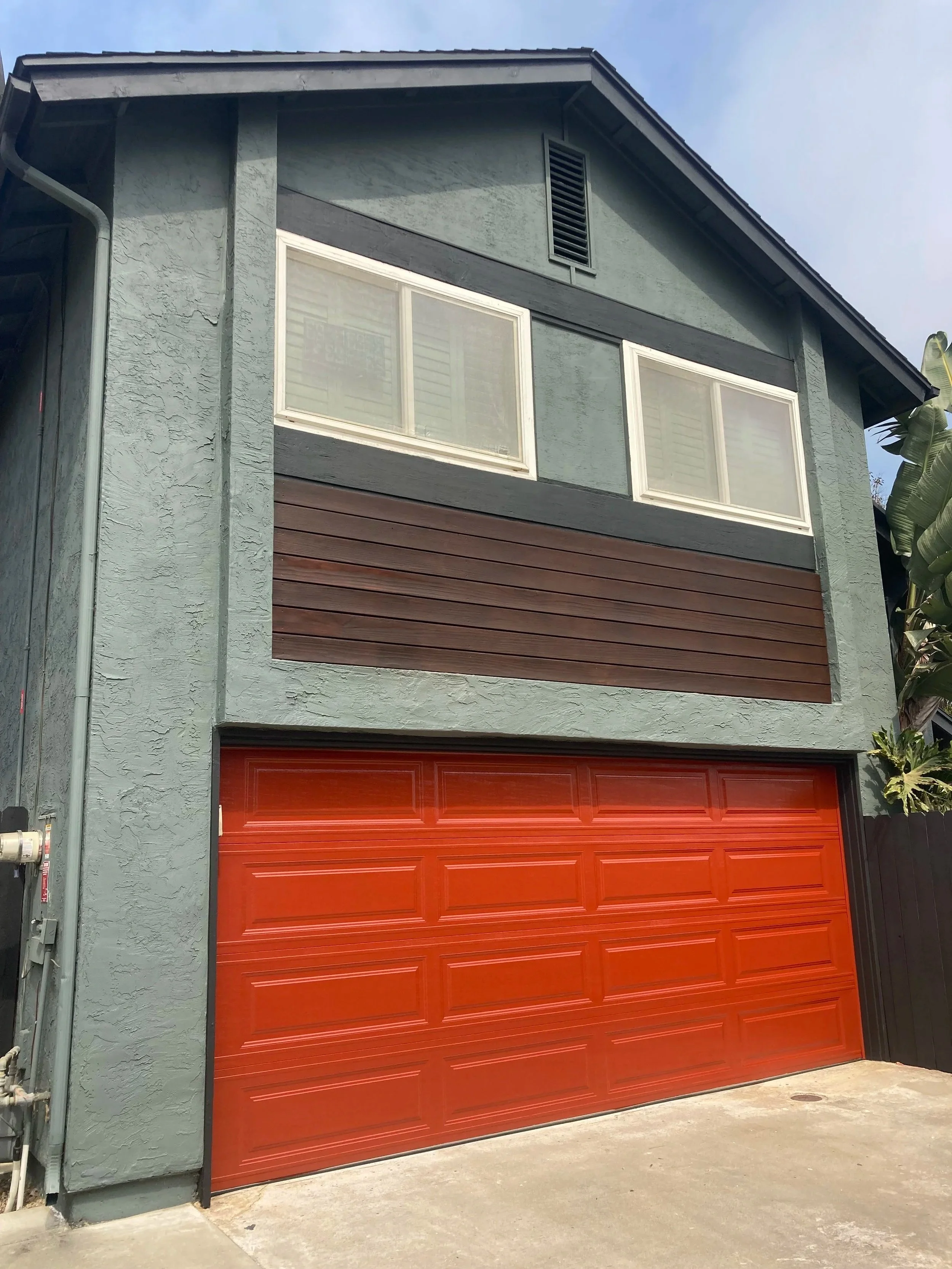 Two-story house with a gray stucco exterior, a red garage door, and a small upper-level window with white frames. The house has a dark trim and a vent near the roof peak.