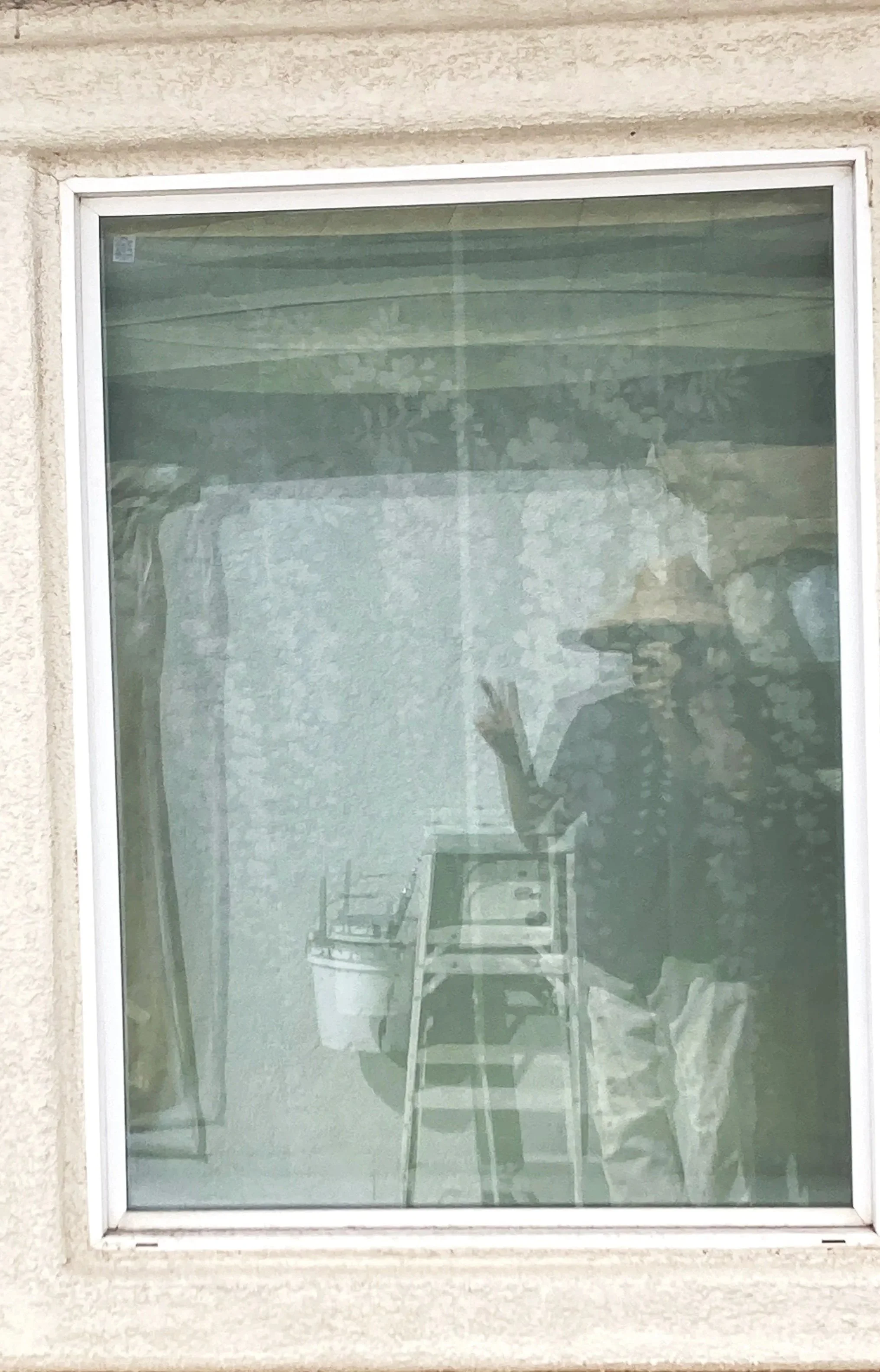 Reflection of person holding a camera or phone, standing outside a window with blinds, with patio furniture visible inside.