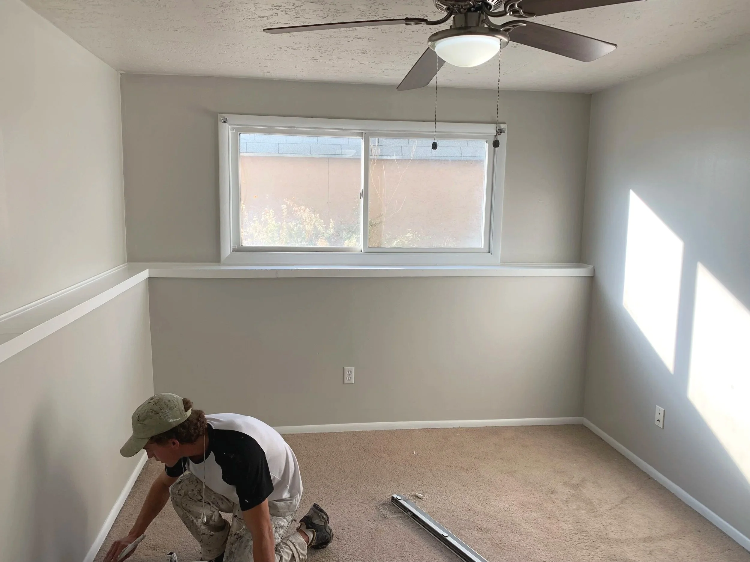 A person kneeling on a beige carpet in a small, empty room with off-white walls, installing baseboards. The person is wearing a beige cap, white and black shirt, and camouflage pants. There is a window on the back wall, a ceiling fan with a light fixture, and an electric outlet visible on the wall.