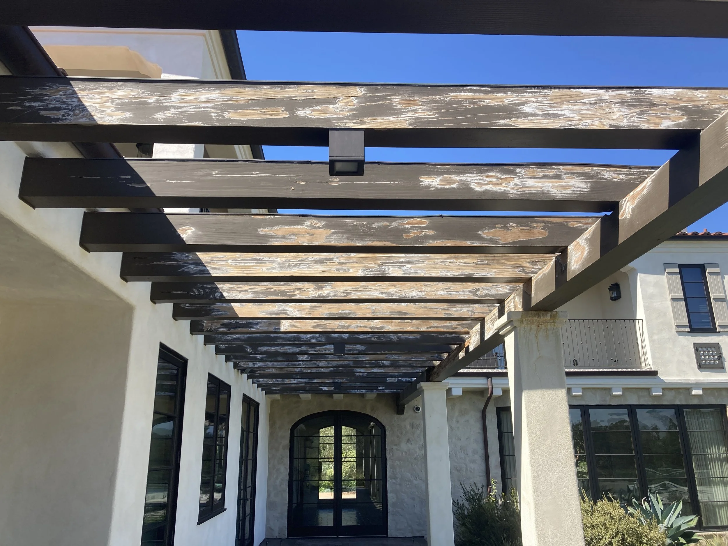Wooden pergola with weathered planks over entrance to a modern house with black-framed windows and white stucco walls, blue sky in background.