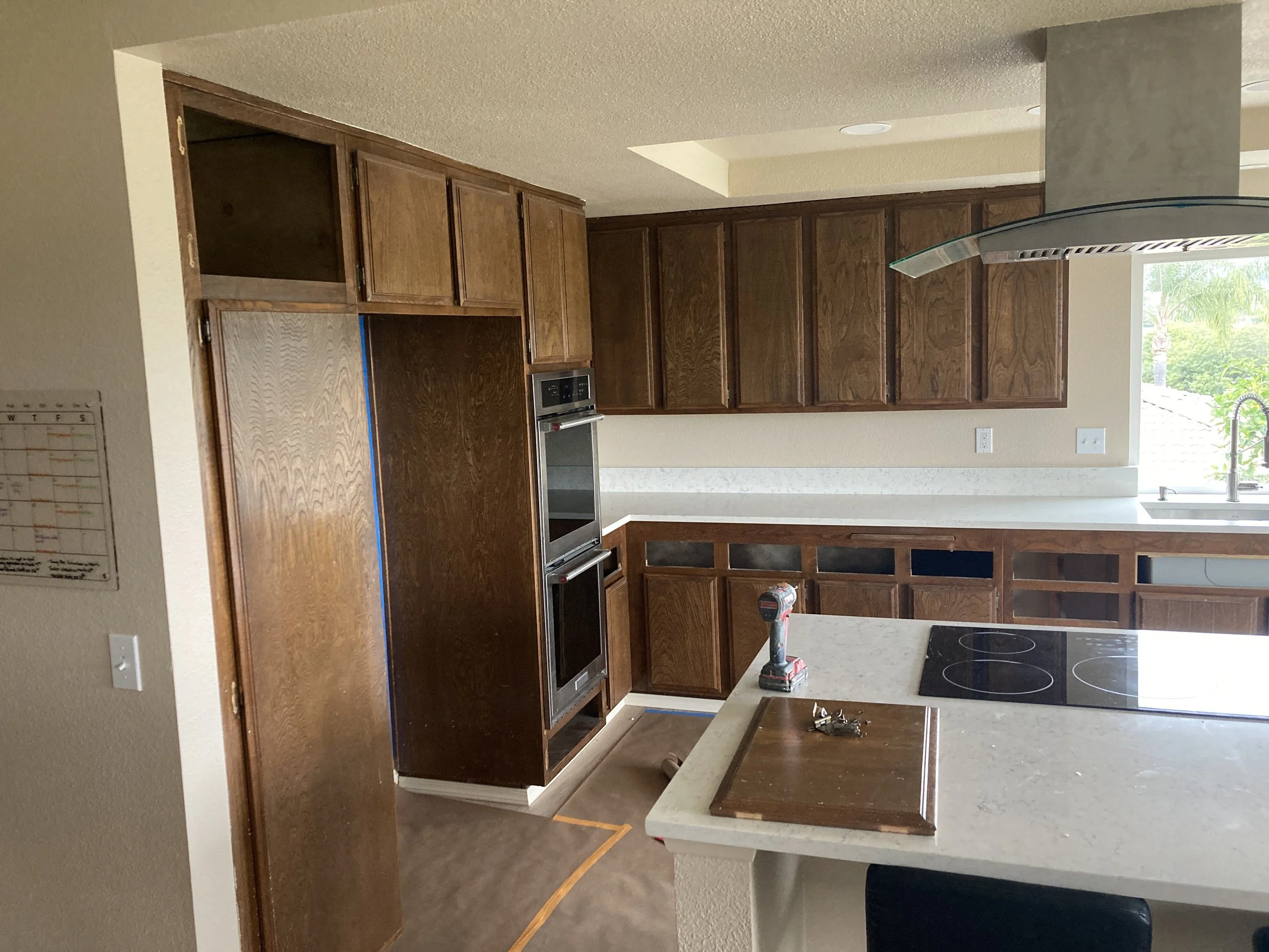 A kitchen with wooden cabinets, a built-in oven, a stove on the island, and a window with greenery outside.