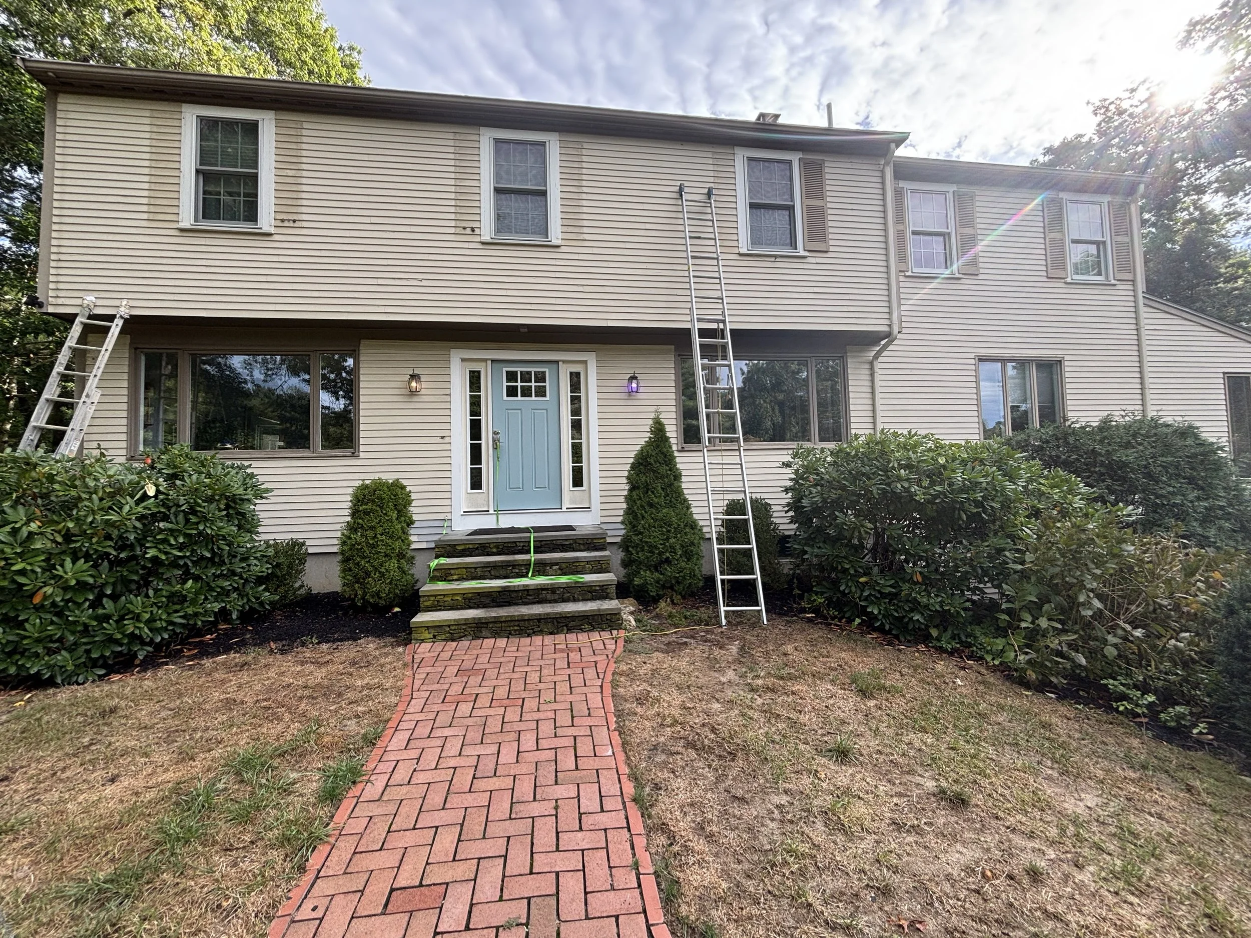 A two-story house with beige siding, six windows, and a blue front door. There is a brick walkway leading to the steps in front of the door. Two ladders are leaning against the house on the left and right sides, with some shrubs and bushes surroundin