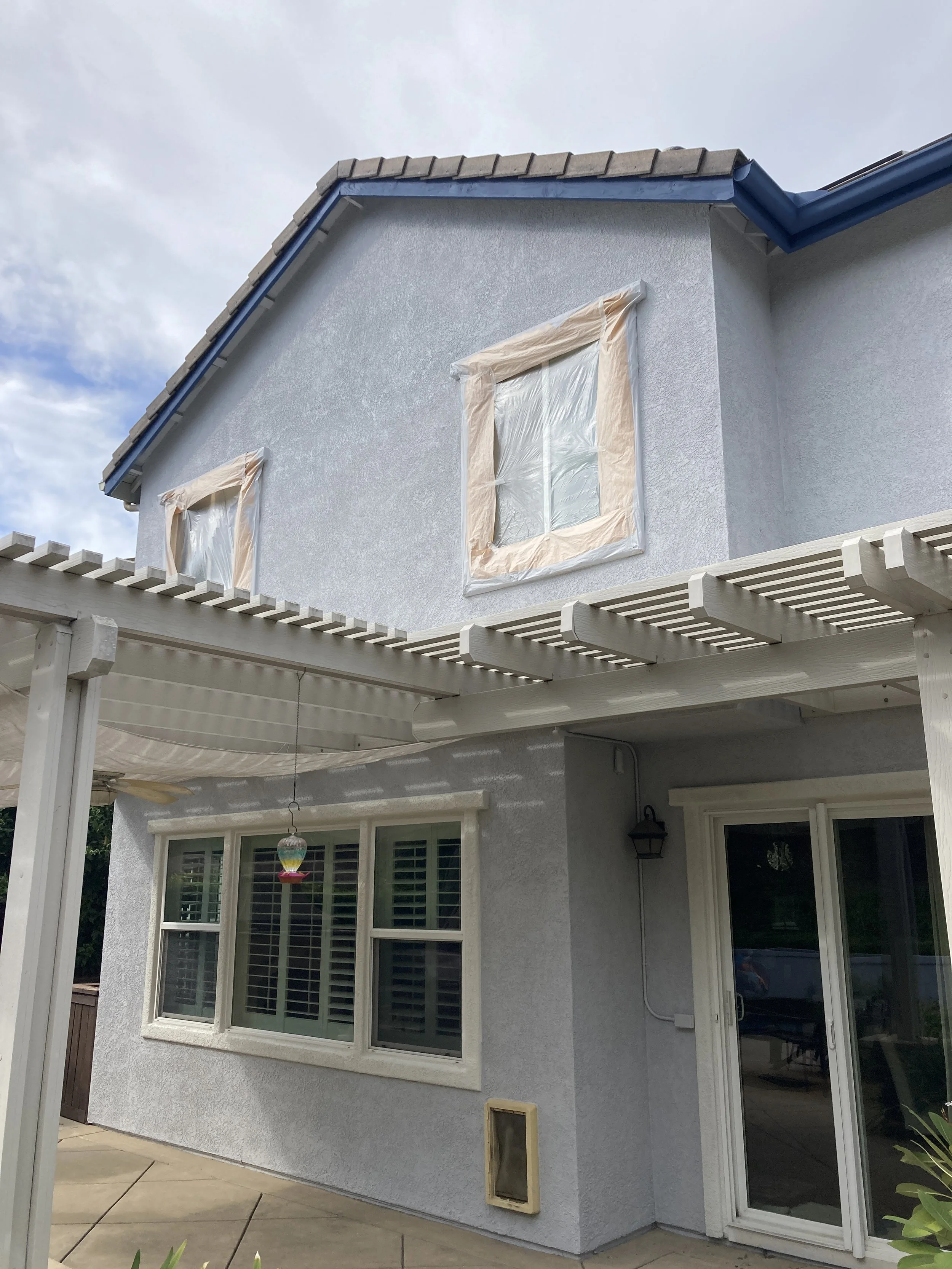 Two windows on a light blue house with paper covering the frames, and a sliding glass door on the ground floor. A pergola with white slats extends from the house, and there is a hanging decoration and a small vent or pet door on the house wall.