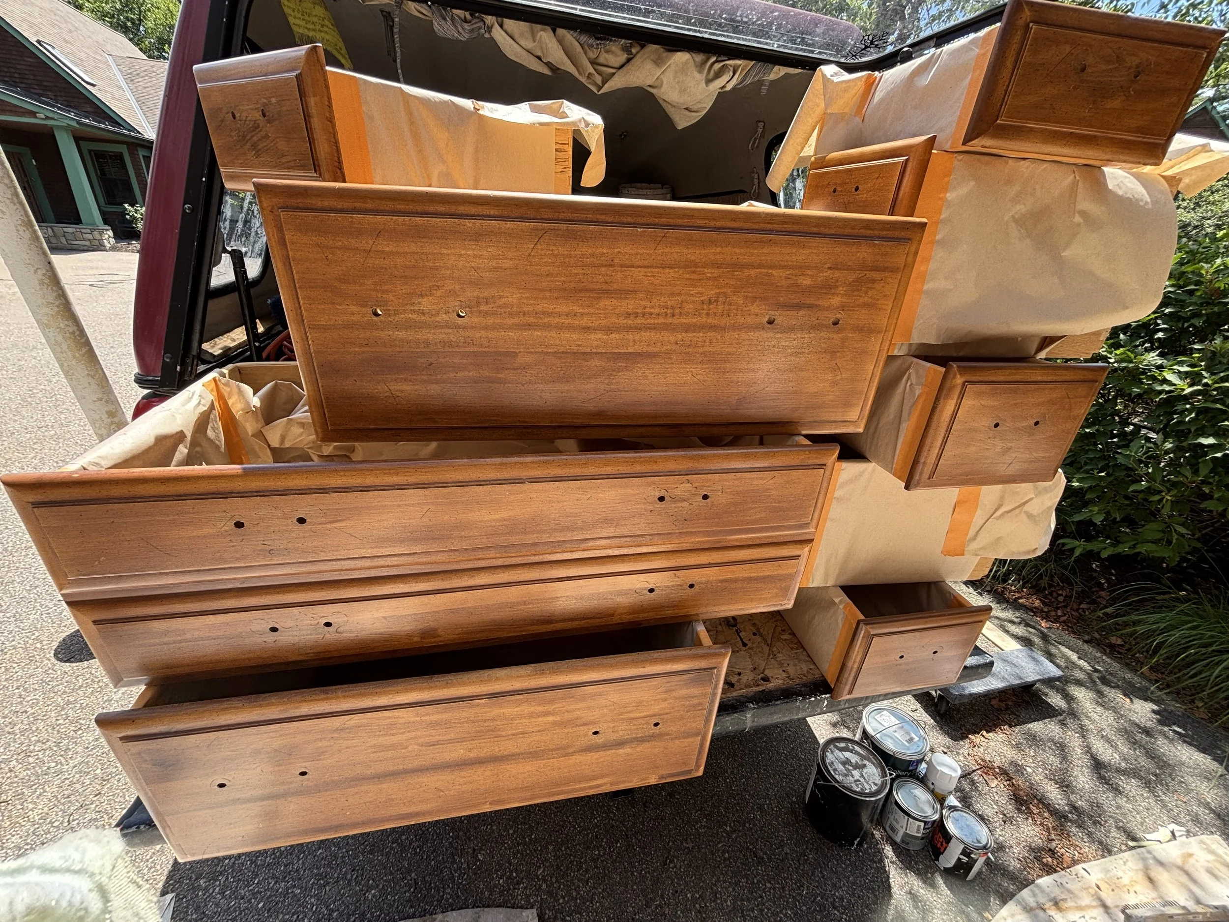Partially painted wooden furniture pieces, including drawers and cabinet doors, are arranged on a truck bed. Several cans of paint are placed on the ground nearby. The furniture appears to be in the process of being painted or restored.