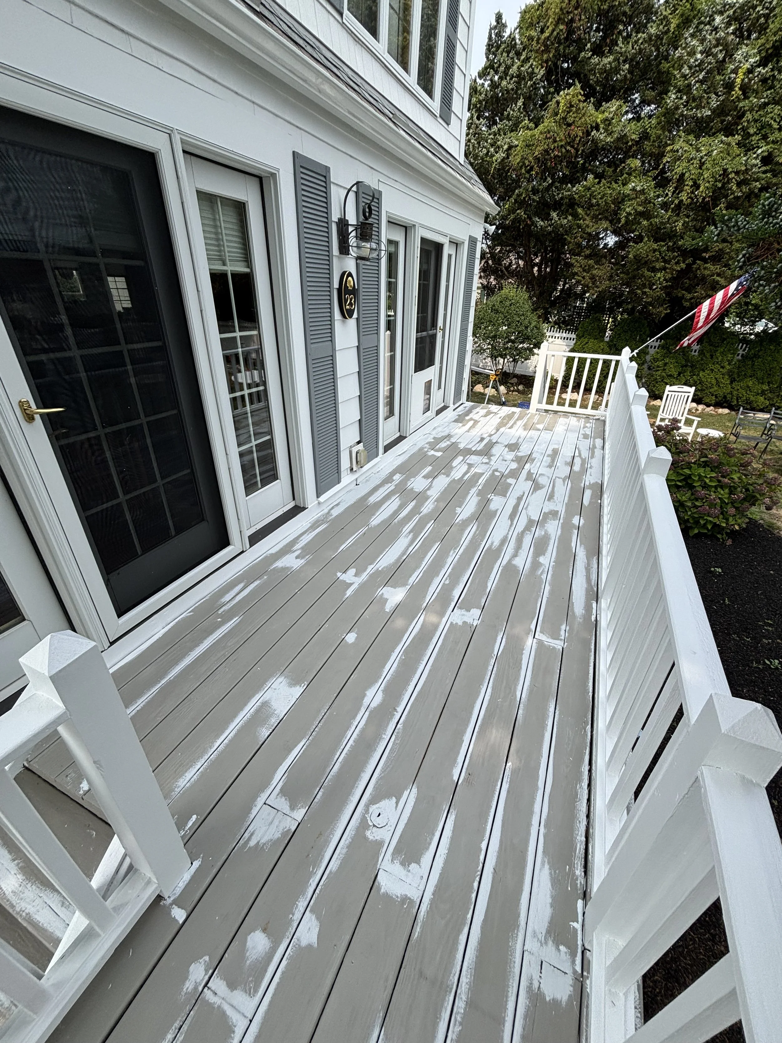 A wooden deck attached to a house with a white railing, showing patches of white paint or primer on the planks, with a door and window on the house's exterior wall, and greenery including trees and shrubs in the background.