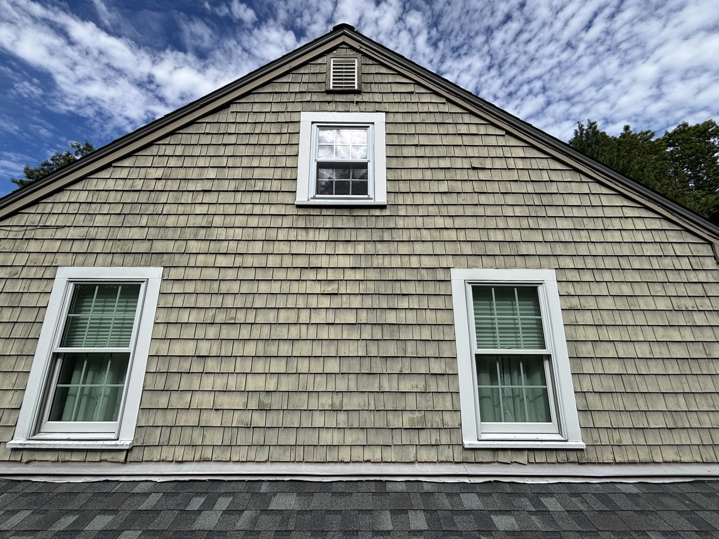 The side of a house with beige shingle siding, three white-framed windows, including one in the gable, and a blue sky with scattered clouds above.