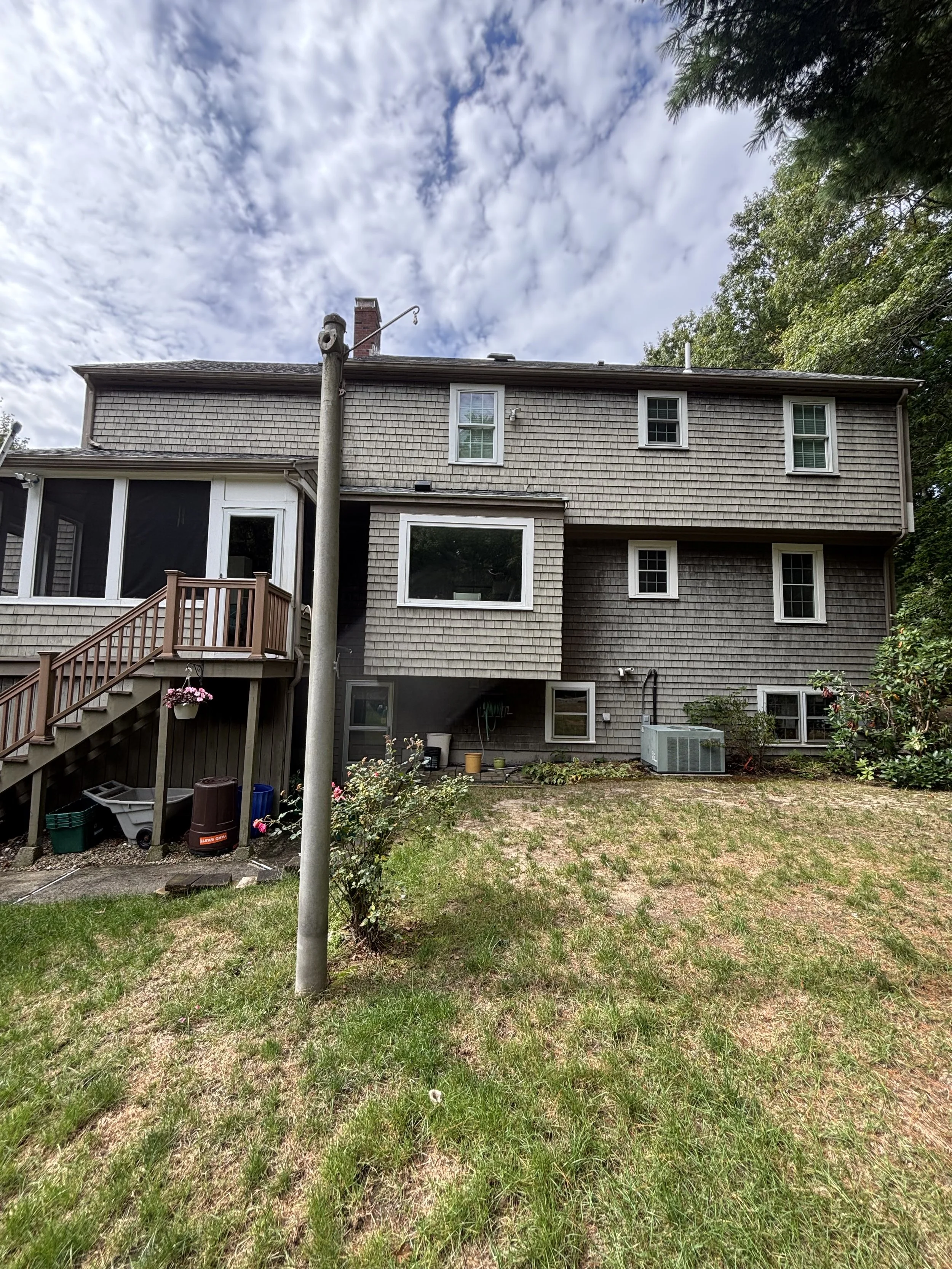 Rear view of a multi-story house with shingle siding, multiple windows, an outdoor staircase, and a small garden area with a grassy lawn and some bushes. A utility pole stands in the foreground, and there is a partly cloudy sky above.