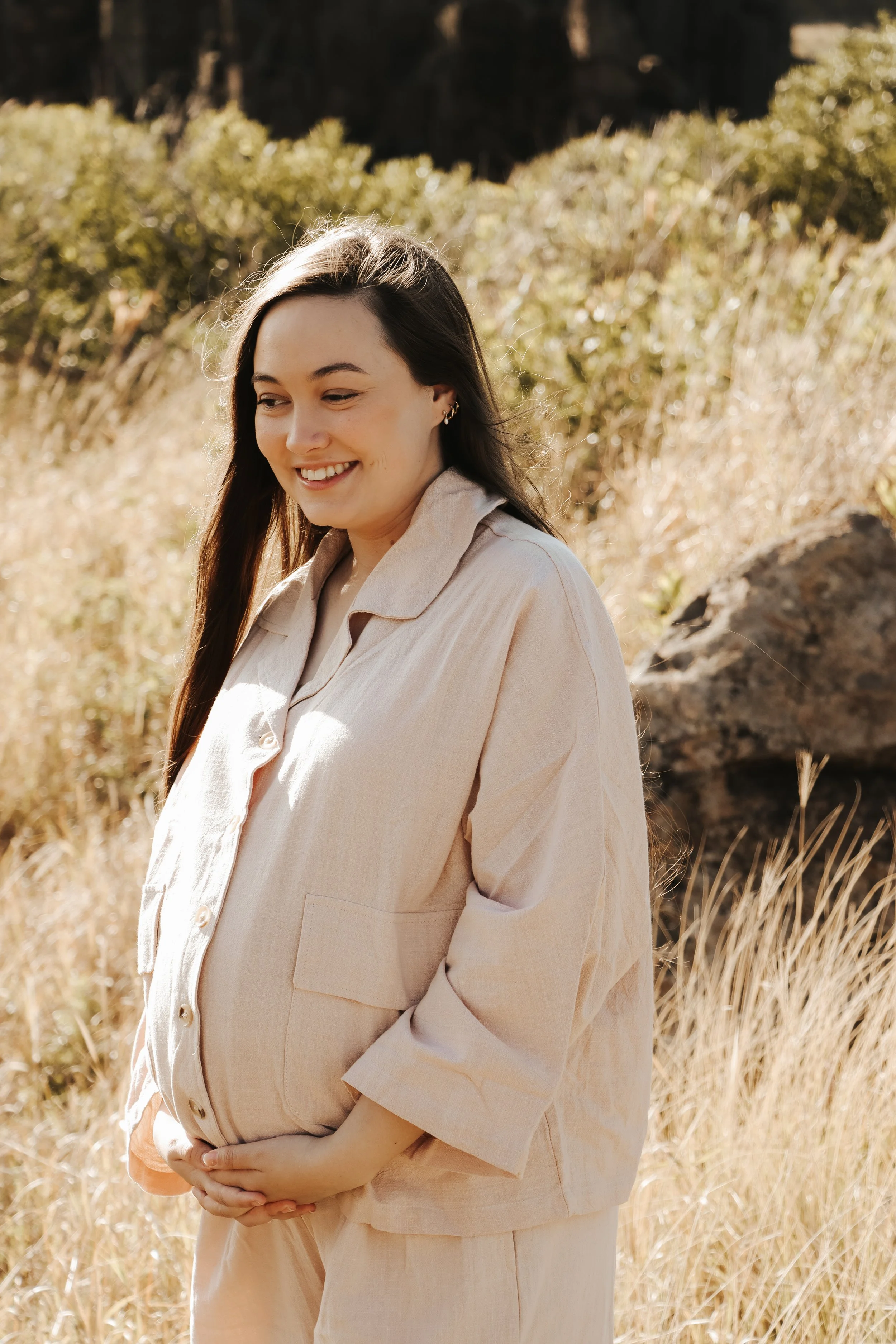A pregnant woman with long dark hair smiling outdoors in a sunny, grassy area with bushes and rocks, wearing a beige outfit. Gentle moment capturing emotional transition into motherhood