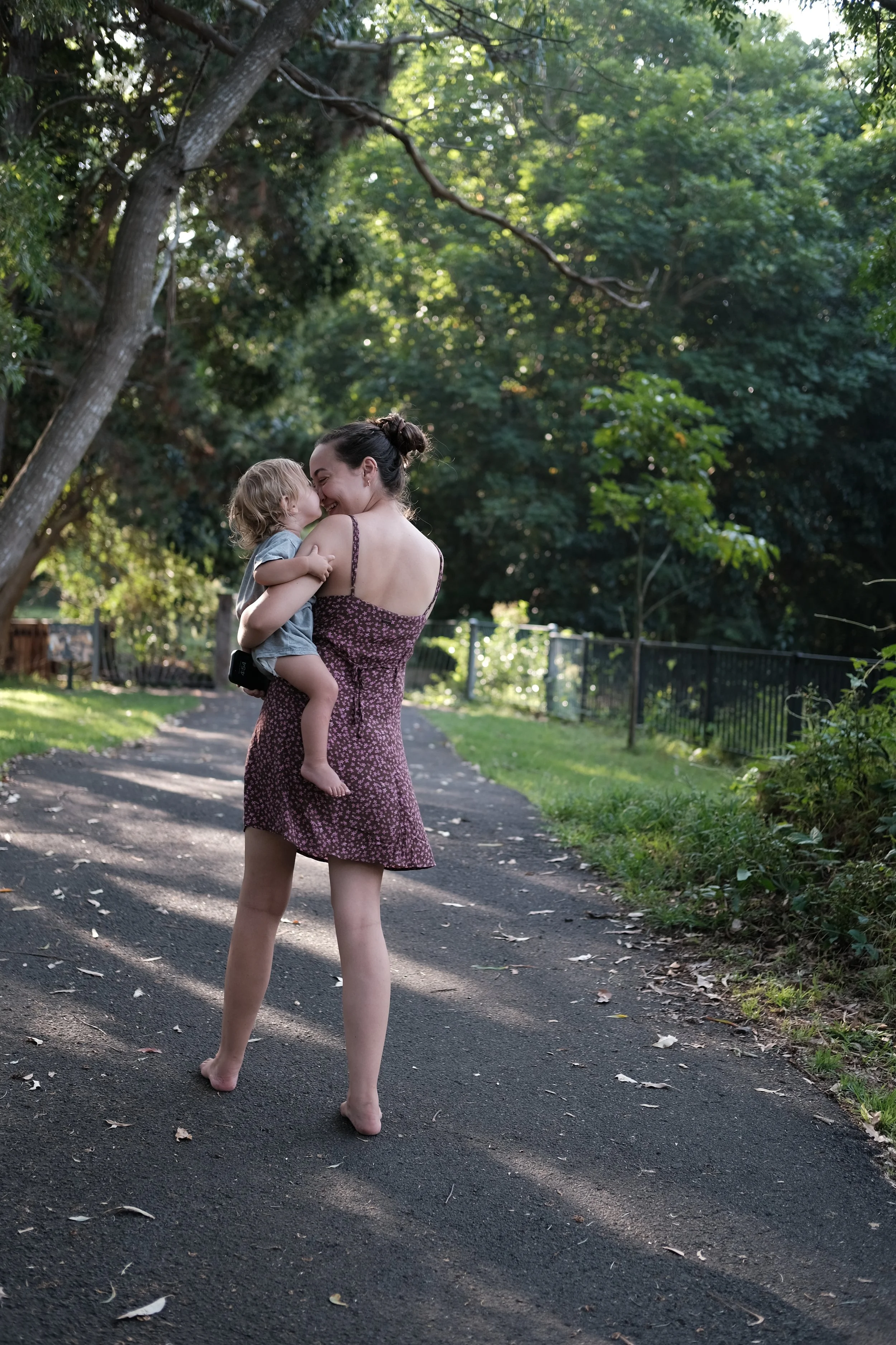 A woman holding a young child on a paved path in a lush green park with trees, smiling and touching noses authentic parenting