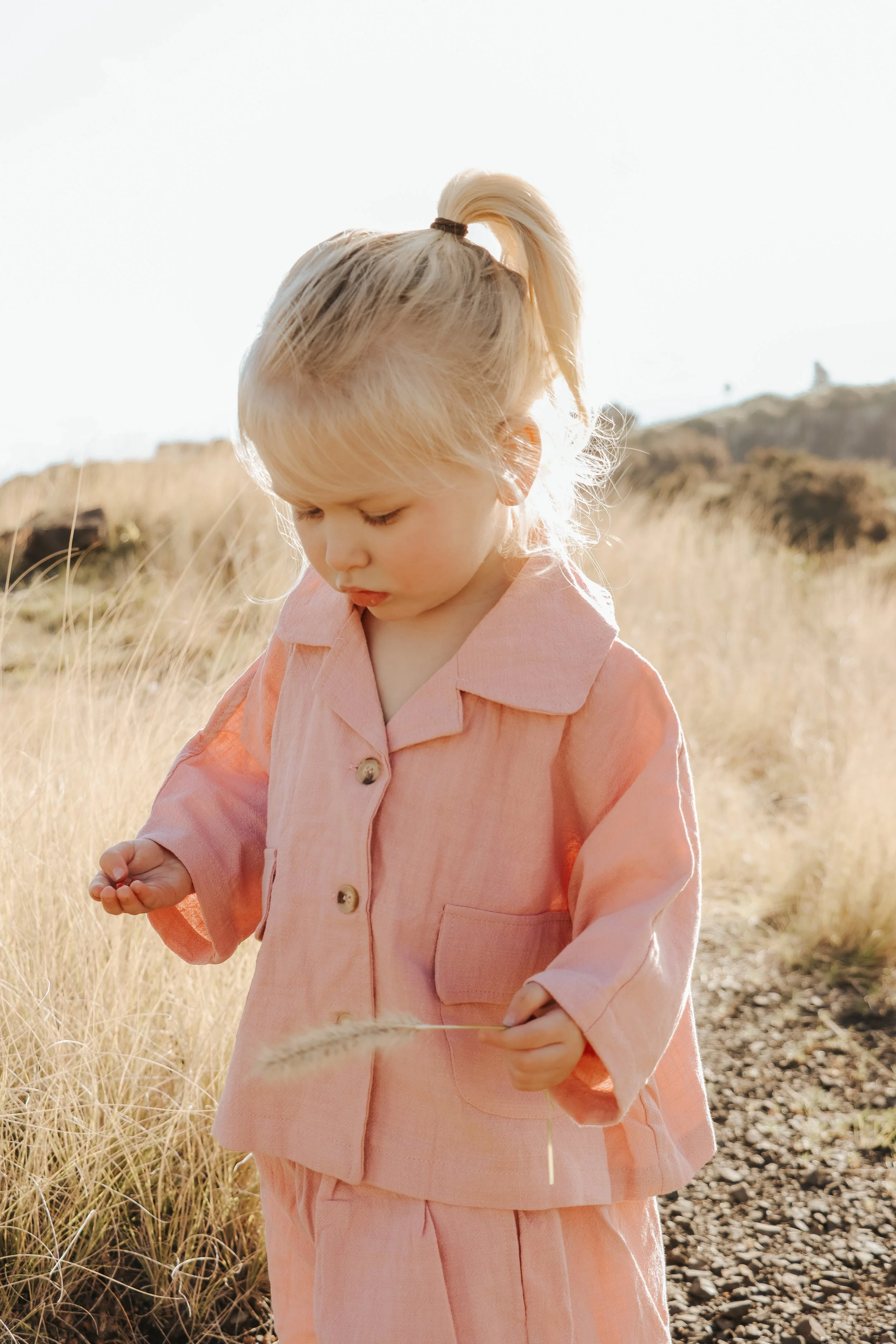 A young girl with blonde hair in a ponytail wearing a peach-colored outfit, standing outdoors in a field of tall, dry grass during sunset or sunrise, looking down at a piece of grass in her hand free outdoor playtime