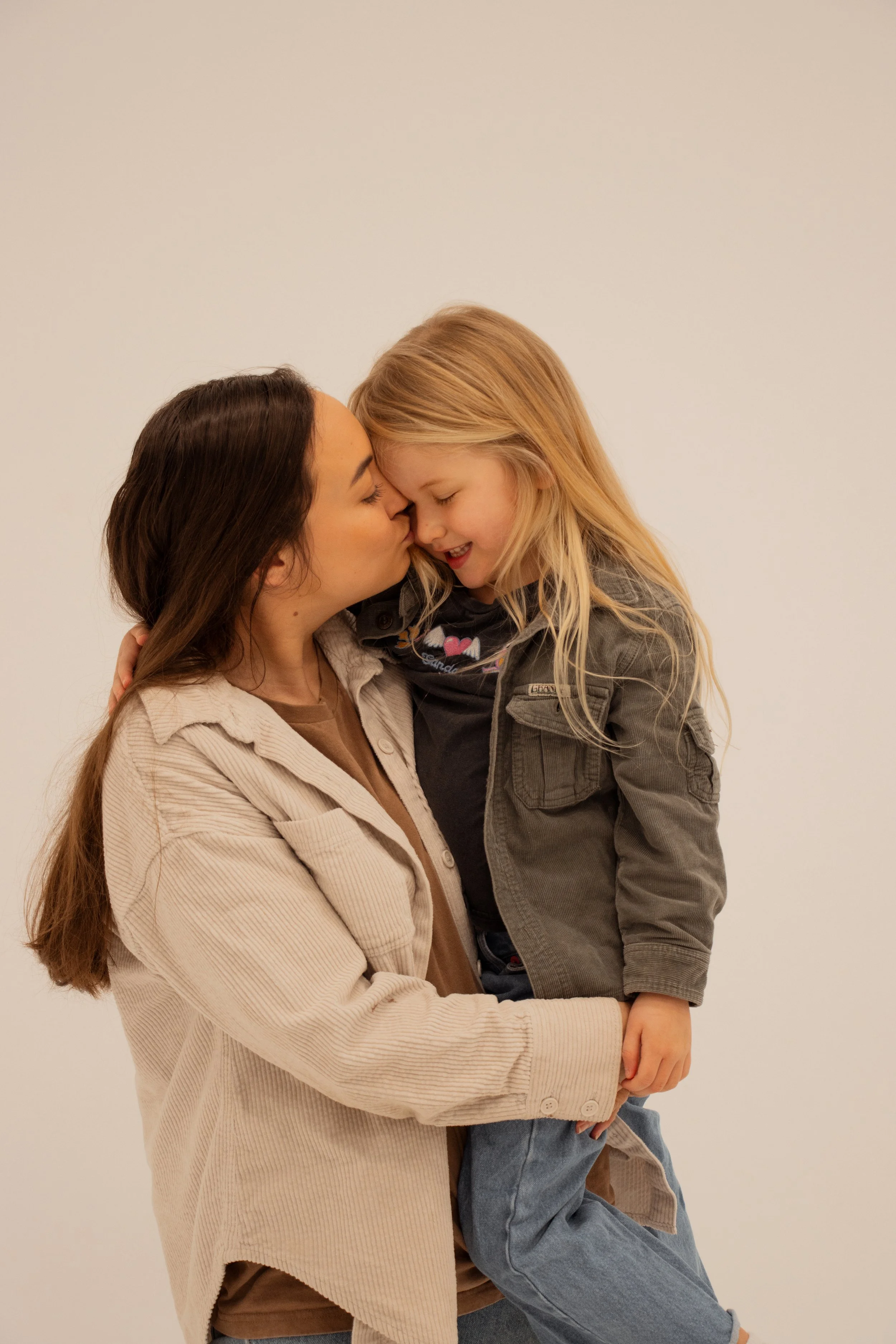 A woman holding a young blonde girl close, touching noses and smiling together against a plain background showing their secure attachment and responsive parenting