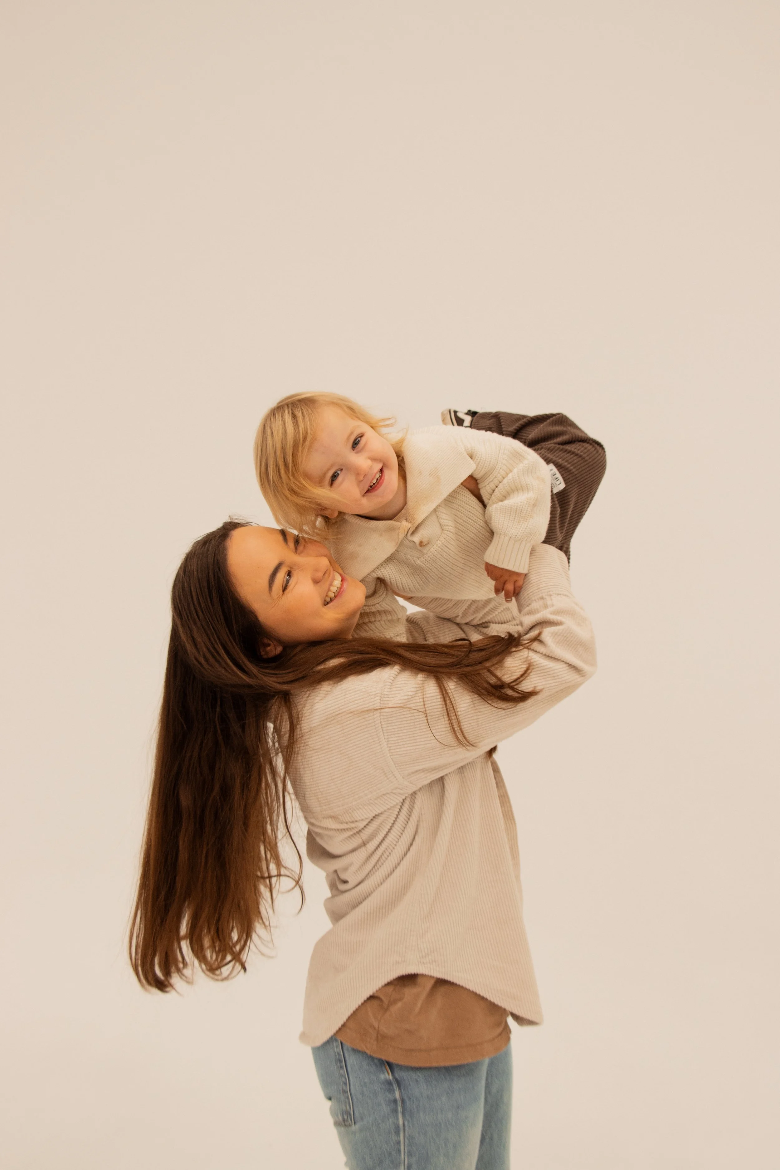 A woman with long brown hair holding a smiling young girl with blonde hair on her shoulders, both wearing neutral-colored sweaters against a plain white wall playing with her children