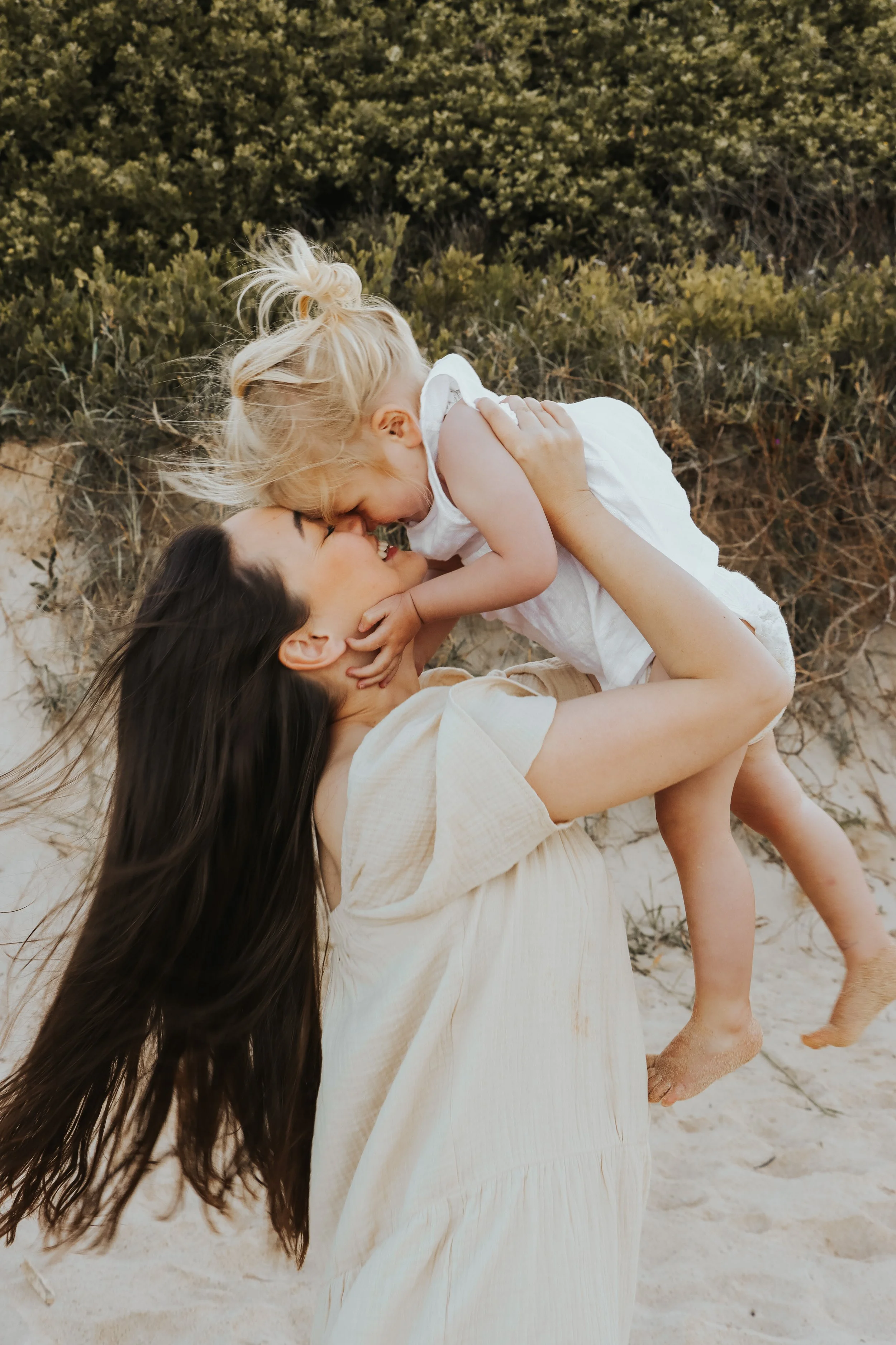 A woman lifting a young girl in the air at the beach, with a background of sand and bushes, both smiling and touching foreheads matrescence and connected parenting