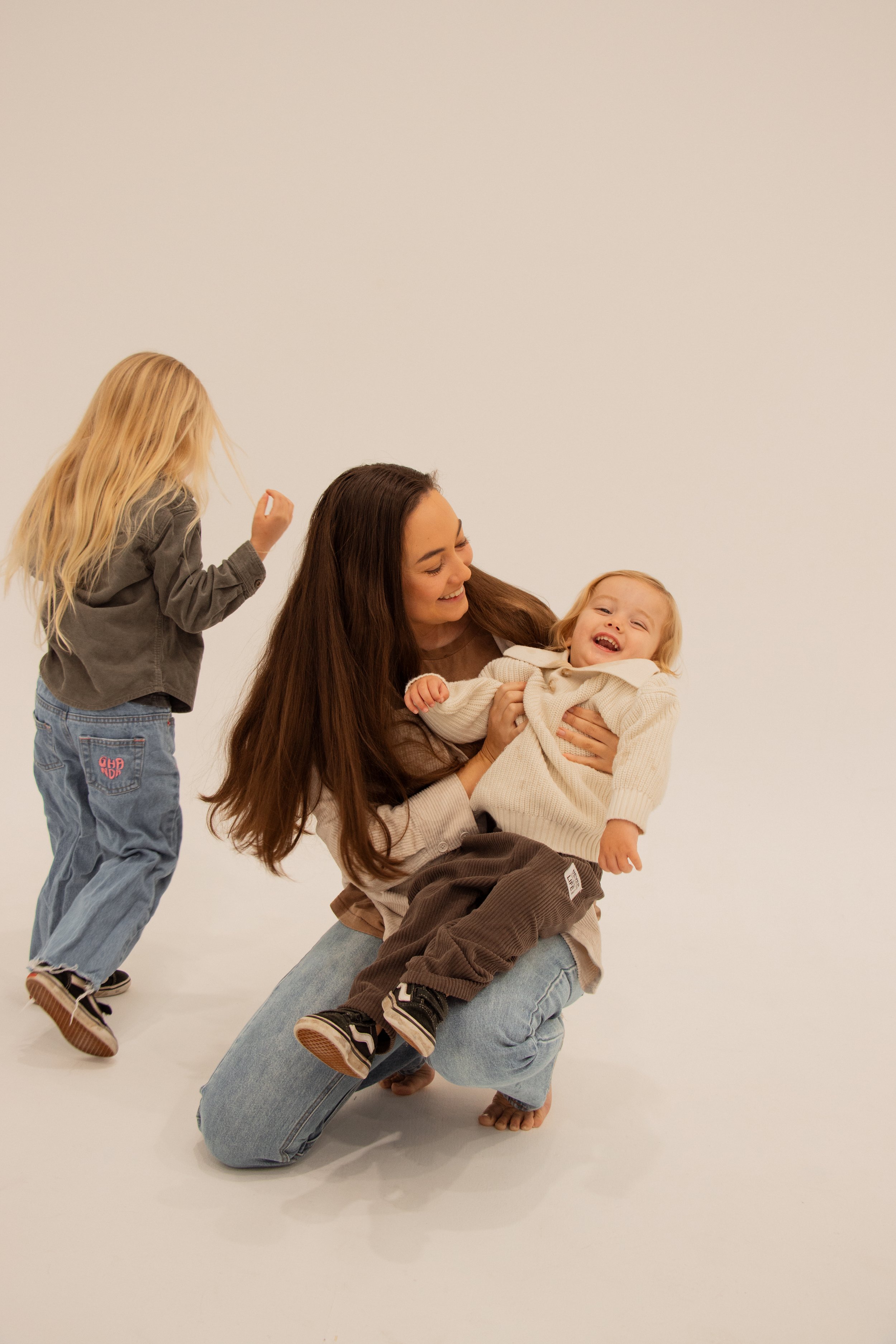 A woman is kneeling on the floor holding a laughing young girl in a cream sweater, while a girl with long blonde hair in a grey hoodie and blue jeans is walking towards them in a white studio with a plain background playful parenting
