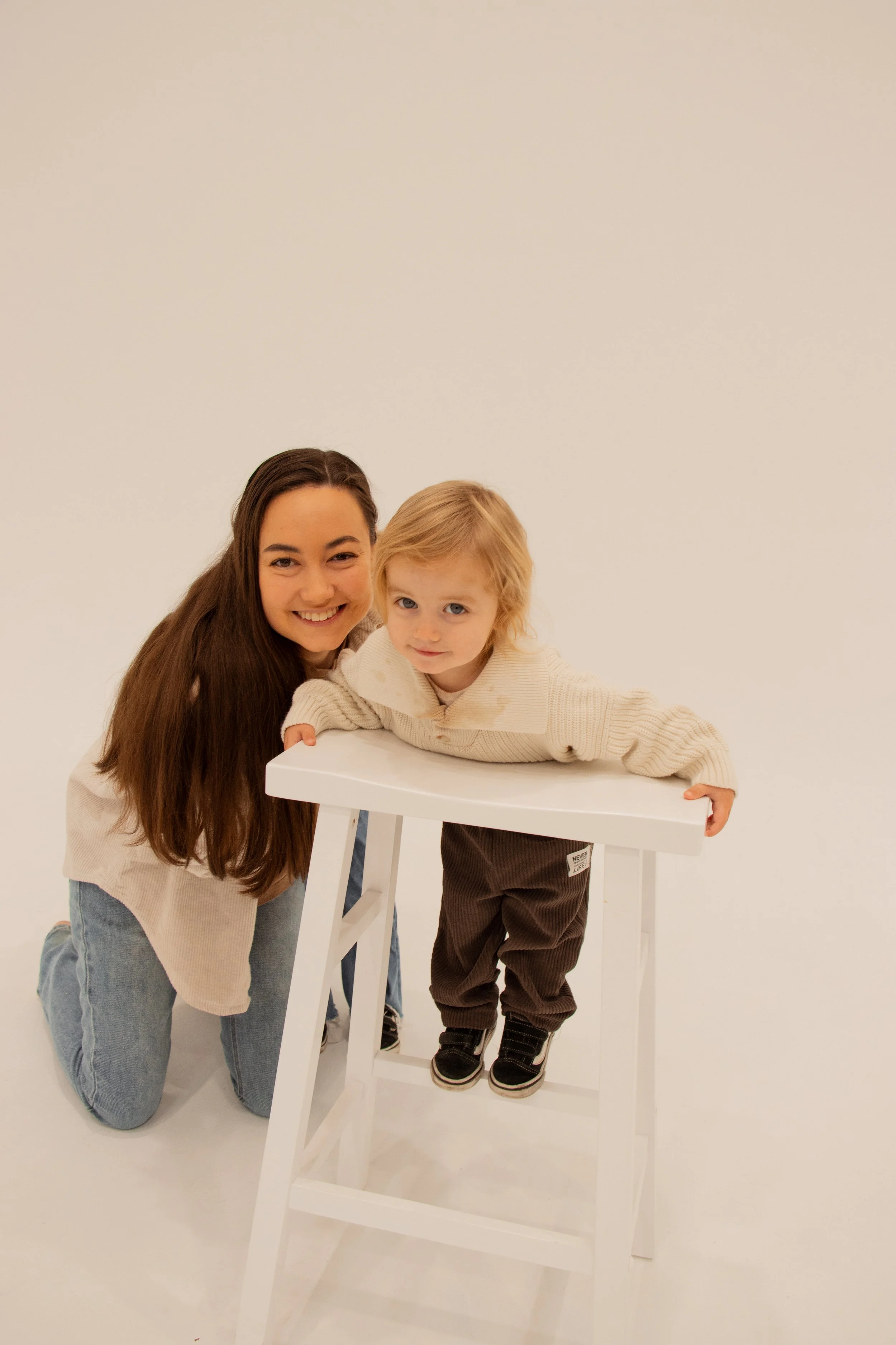 A woman kneeling beside a young boy leaning on a white table against a plain white background playing on the floor with relaxed connection