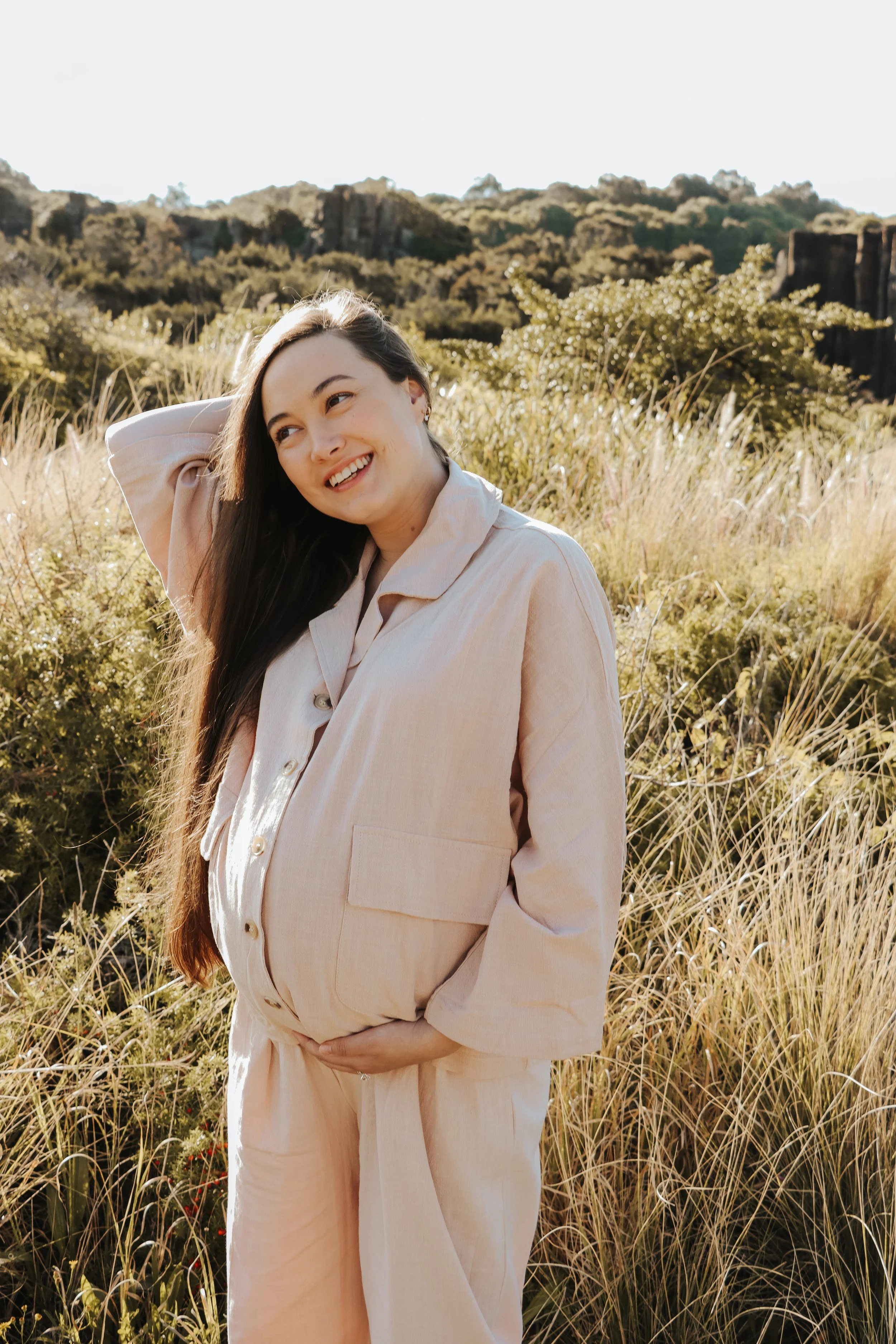 Sheridan from Early Nurturing pregnant and smiling with long dark hair, wearing a light beige outfit, standing outdoors in a field of tall grass and plants with a scenic background of trees and rocky formations, during golden hour.