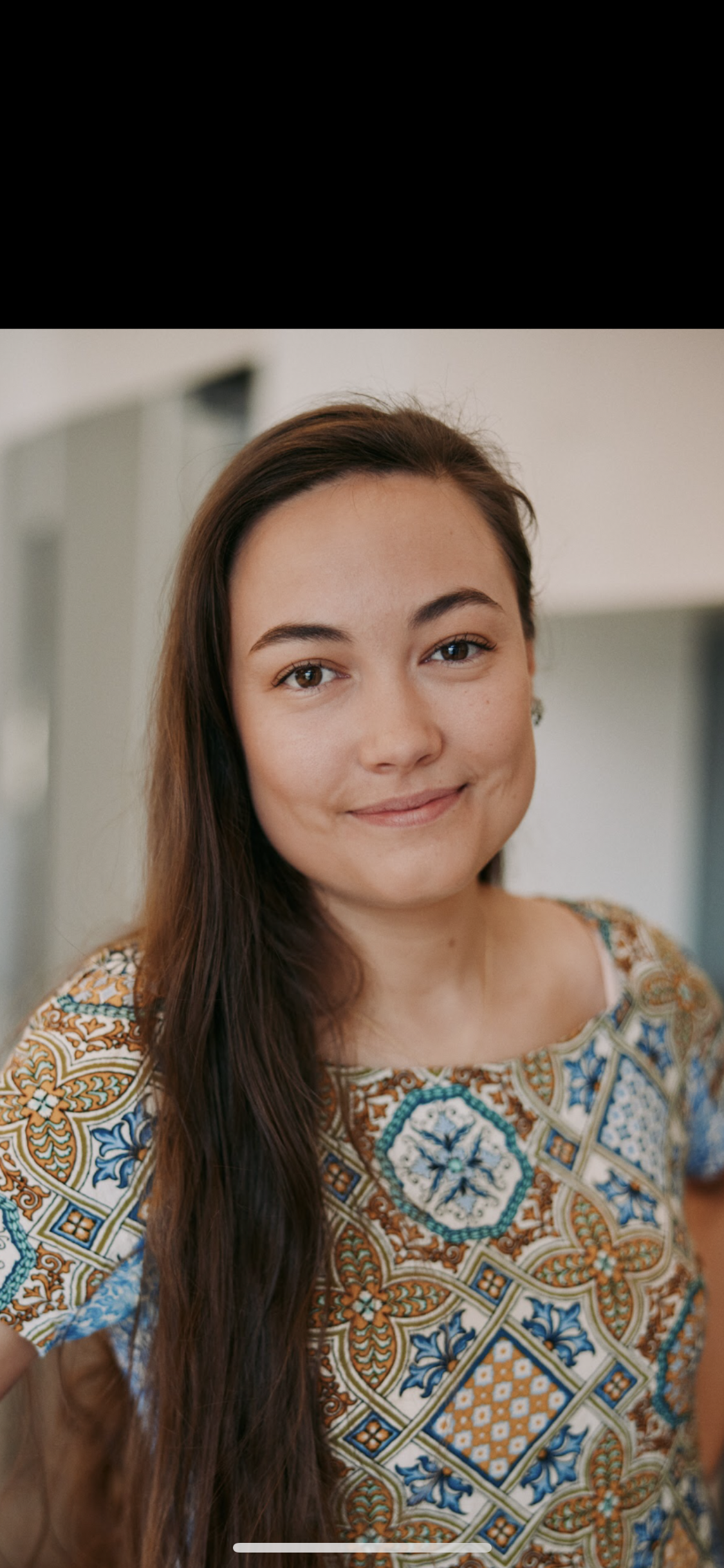 Close-up of a young woman, Sheridan from Early Nurturing, Sleep and early parenting specialist with long brown hair, smiling softly, wearing a colorful patterned top, standing indoors with blurred background.