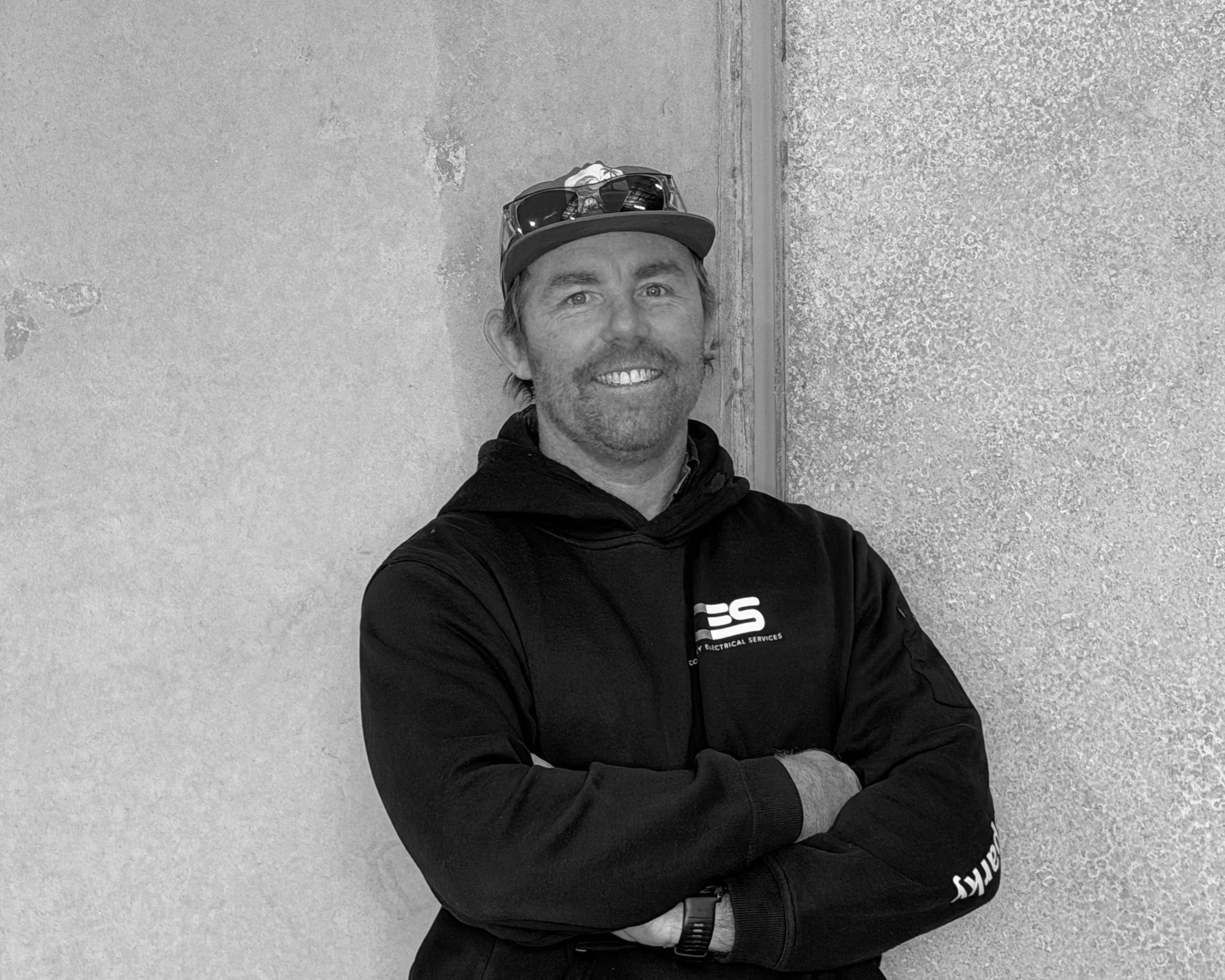 A man smiling and wearing a baseball cap with sunglasses on top, standing against a textured wall, dressed in black clothing with a logo on his jacket.