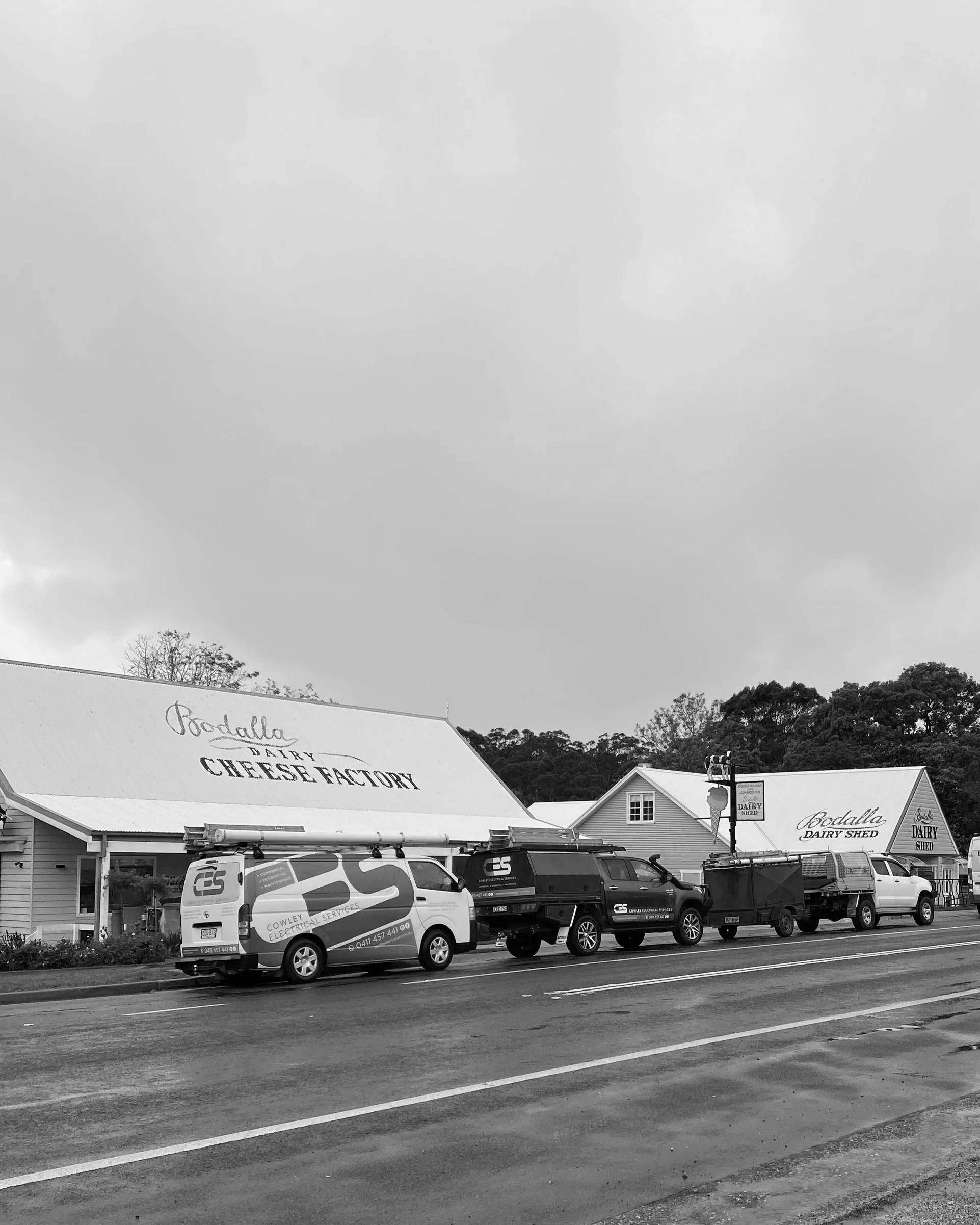 A row of vehicles parked in front of a building with a large sign reading 'Bodalla Dairy Shed' on the roof, with trees and a cloudy sky in the background.