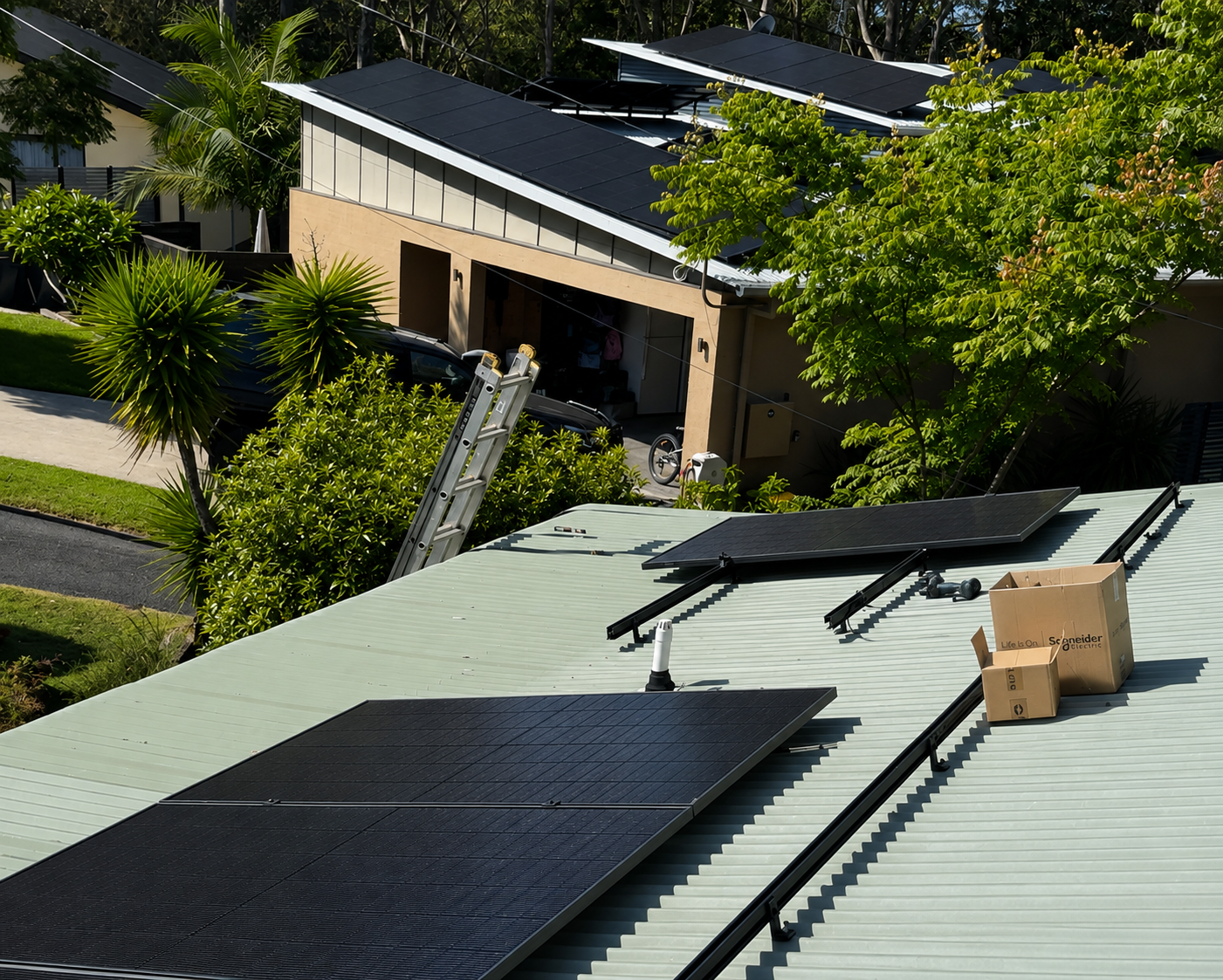 A rooftop with solar panels in the process of installation, a cardboard box, ladder, tools, and trees with green foliage surrounding a modern house with a garage and driveway.