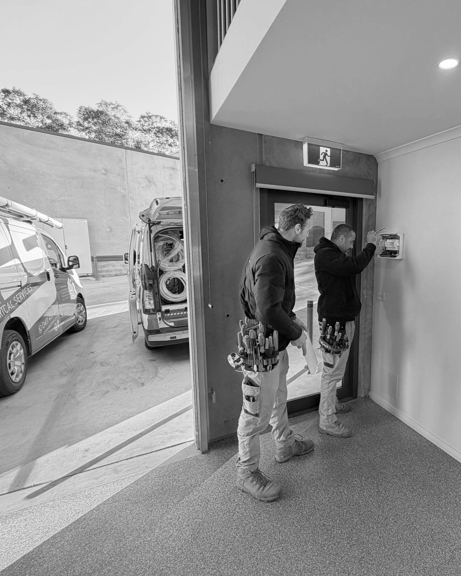 Two workers with tool belts standing near an entry door, one about to enter and the other already inside, with vehicles and equipment outside.