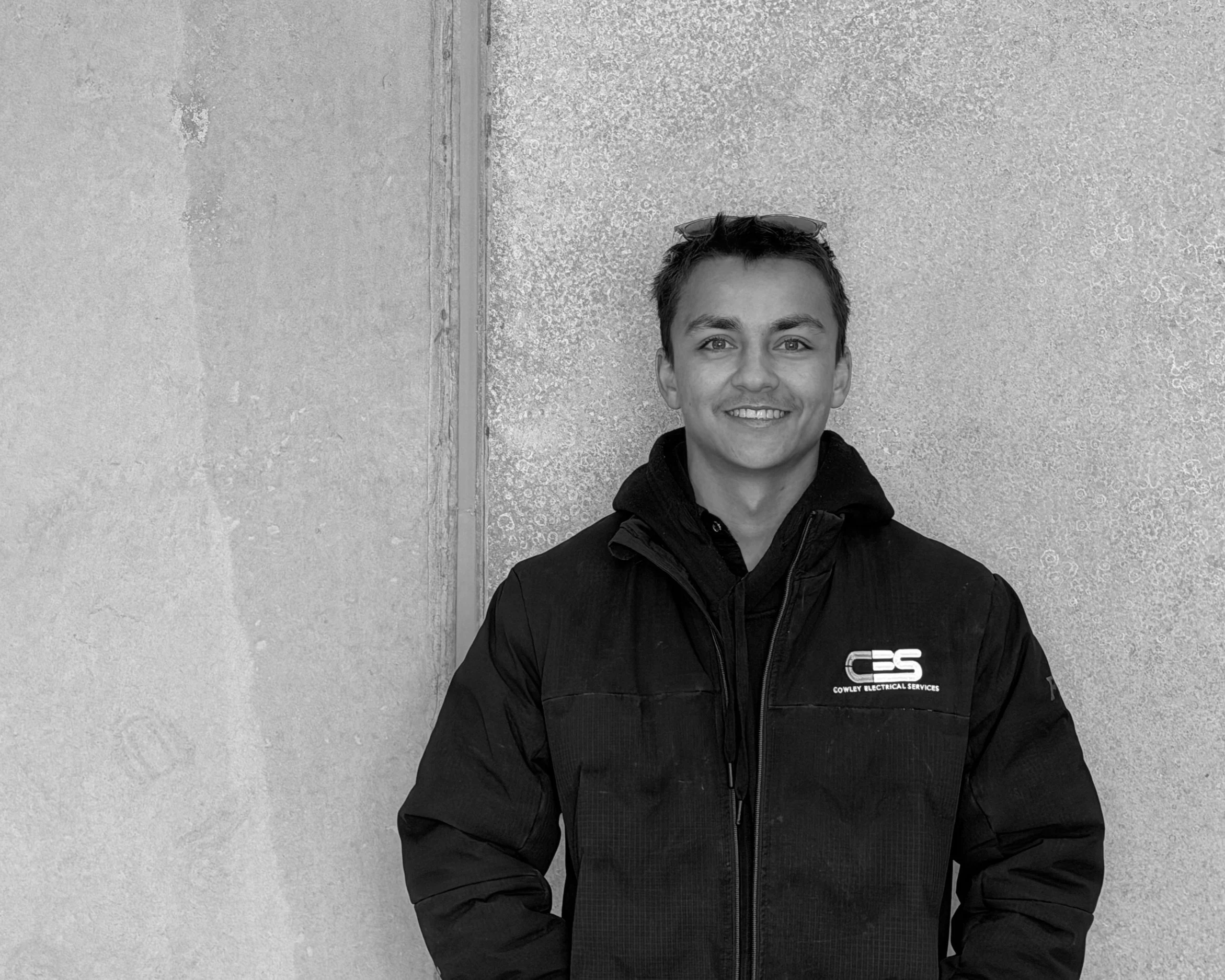 Young man smiling, wearing a black jacket with a logo, standing in front of a textured wall, sunglasses on his head.