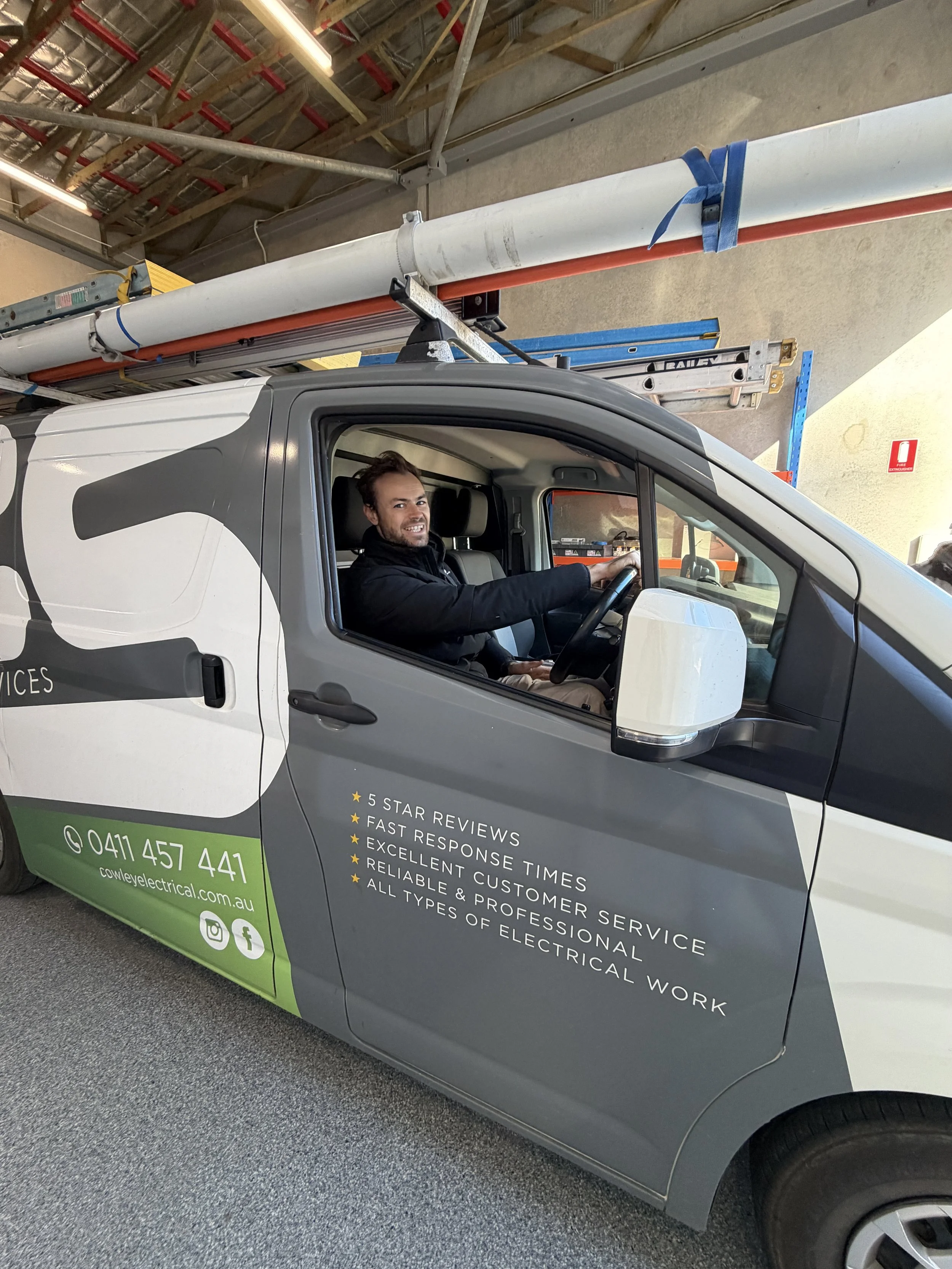 A man sitting in the driver's seat of a service van, smiling at the camera. The van has advertising for electrical services on the side, including a phone number and website. The van is parked indoors, with shelving, a ladder, and construction materials visible in the background.
