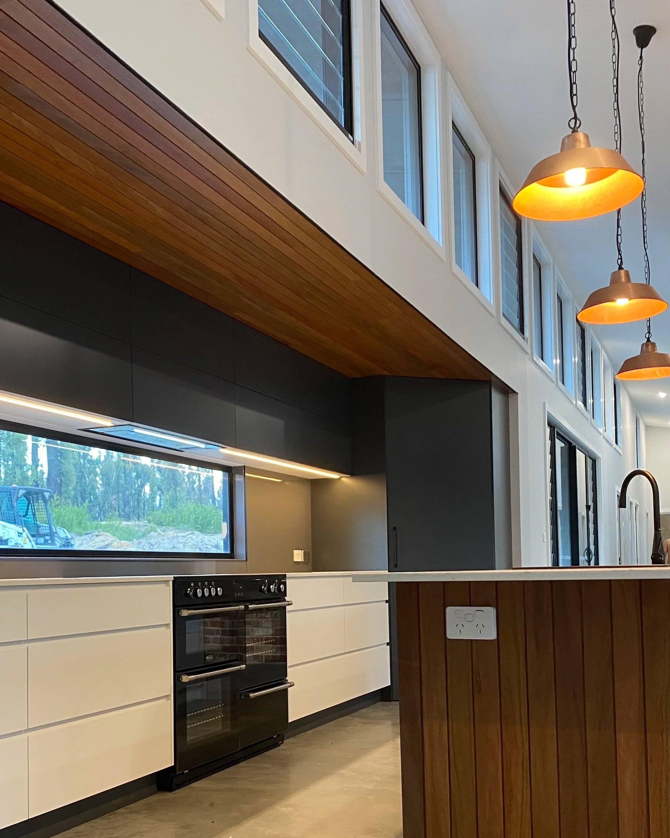 Modern kitchen with white cabinets, black oven, wooden ceiling, orange pendant lights, large window showing outdoor greenery.