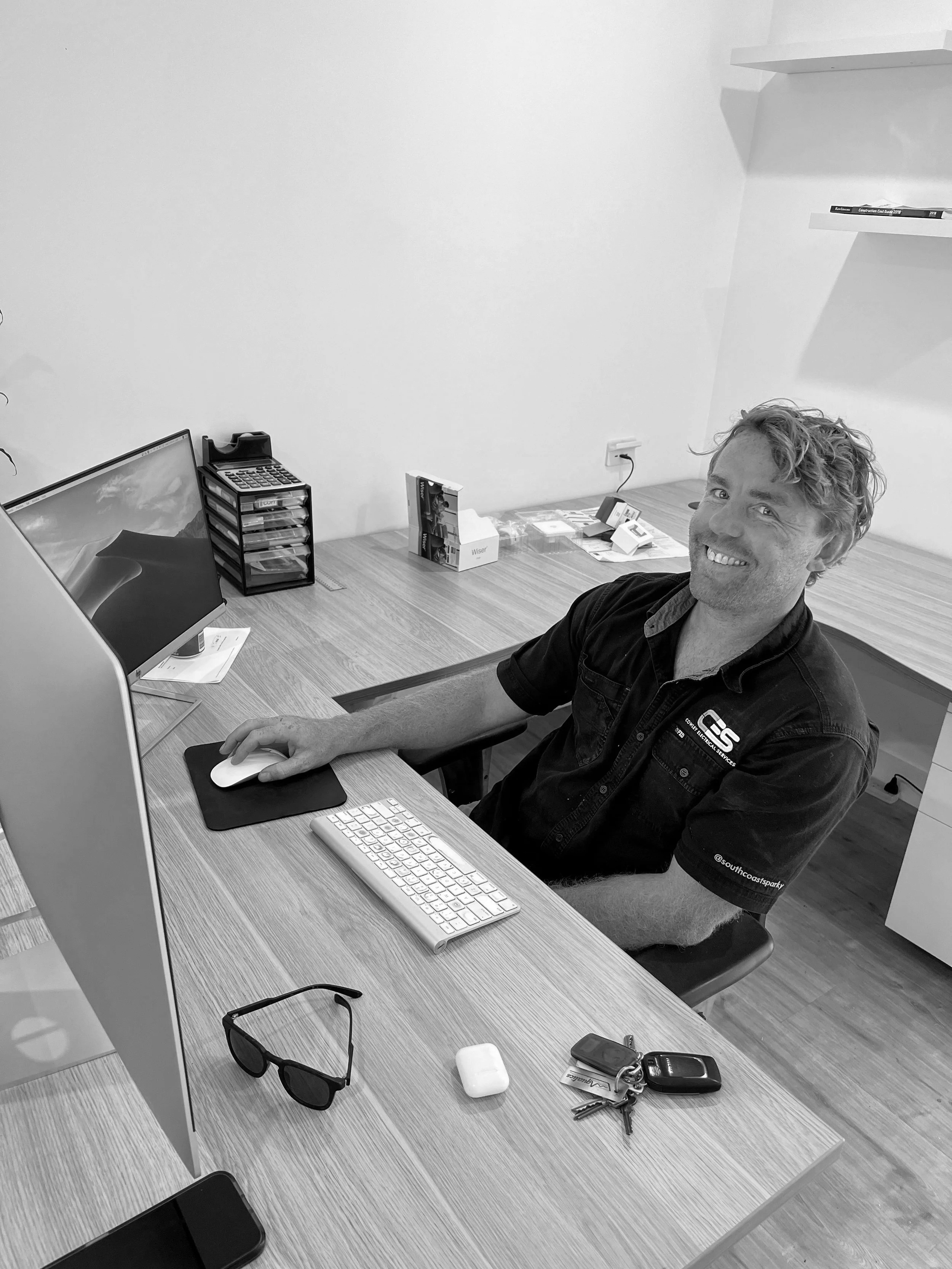 A man sitting at a desk with a computer monitor, keyboard, mouse, sunglasses, keys, and a pair of sunglasses on the desk. There are magazines and other office supplies, with a second shelf visible on the wall.