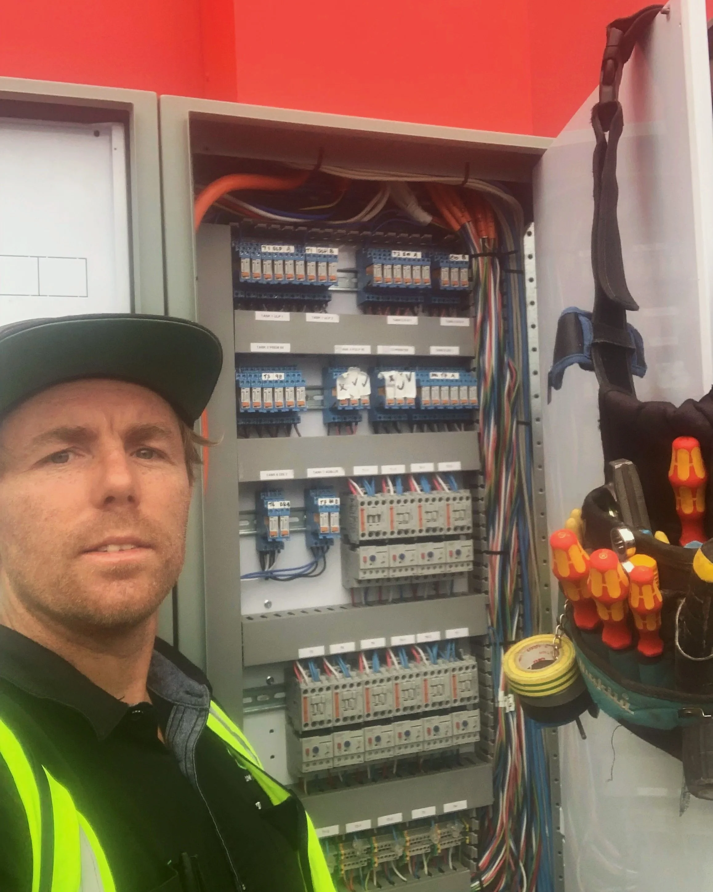 A man wearing a black cap and high-visibility safety vest taking a selfie in front of an open electrical control panel with various wires, circuit breakers, and tools hanging on the side.