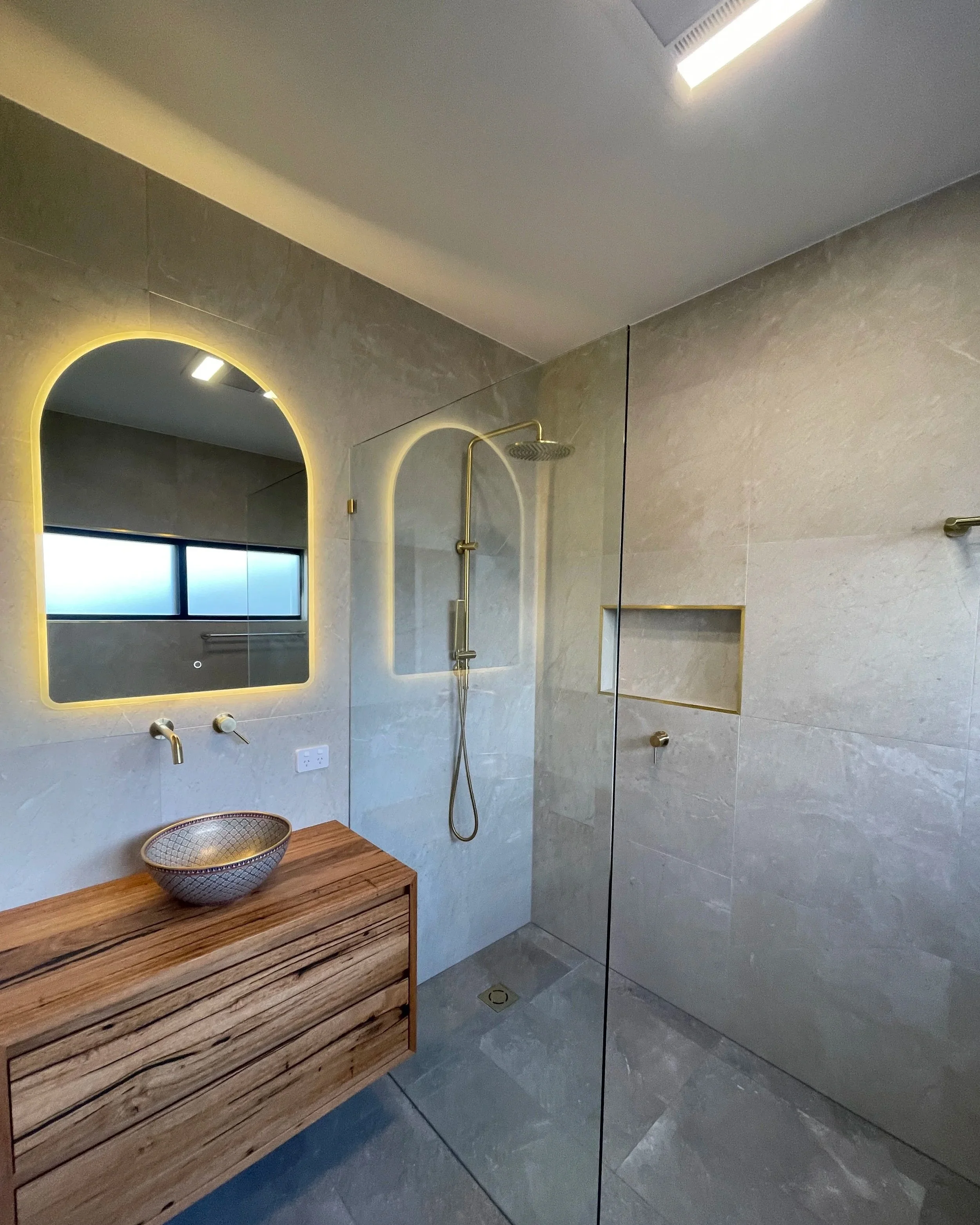 Modern bathroom with beige tiled walls, a round mirror with LED lighting, a showerhead with a handheld spray, and a wooden vanity with a decorative bowl.