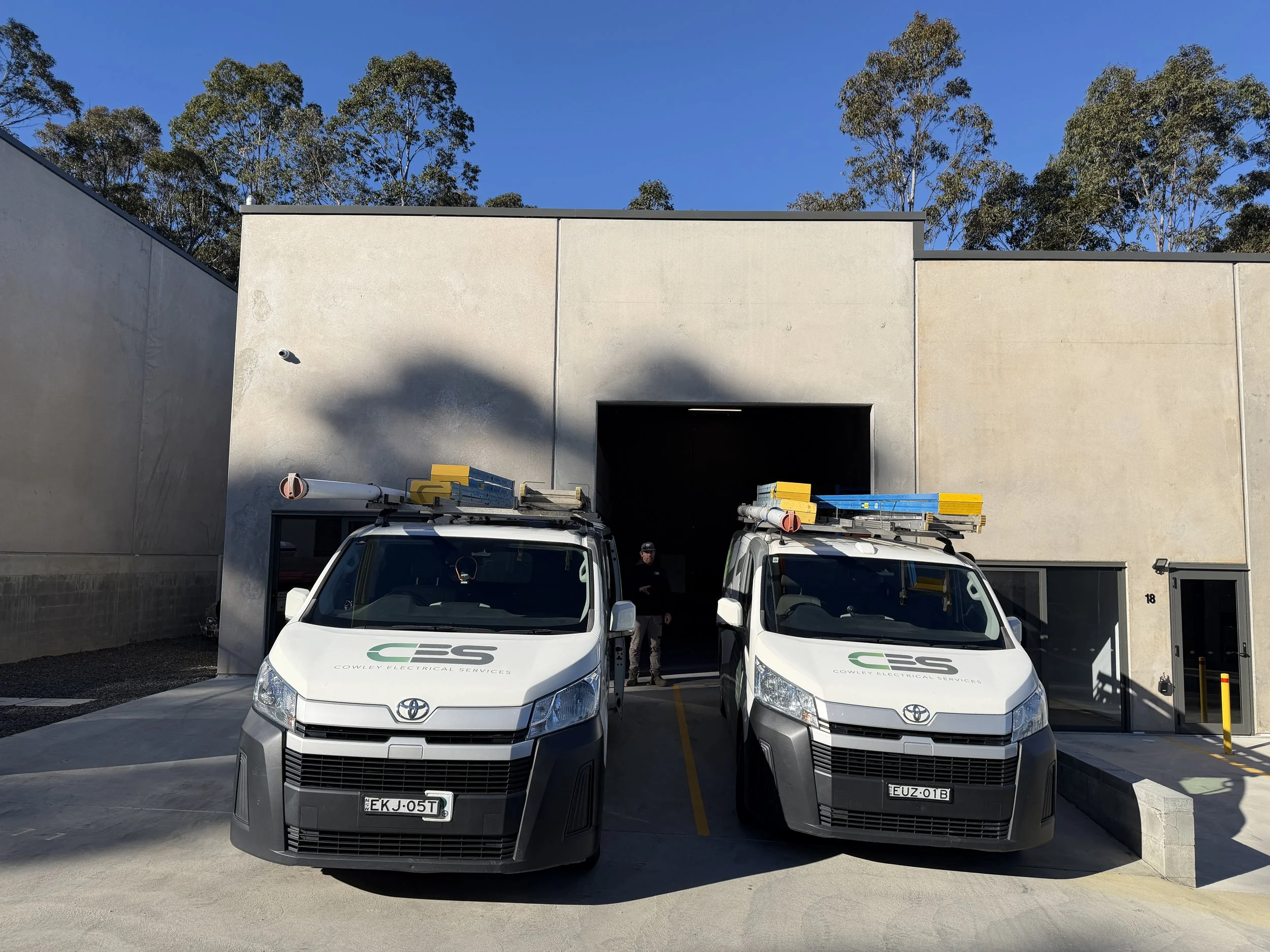Front view of two white company vans with ladders on top parked in front of a beige building, with trees and a blue sky in the background.