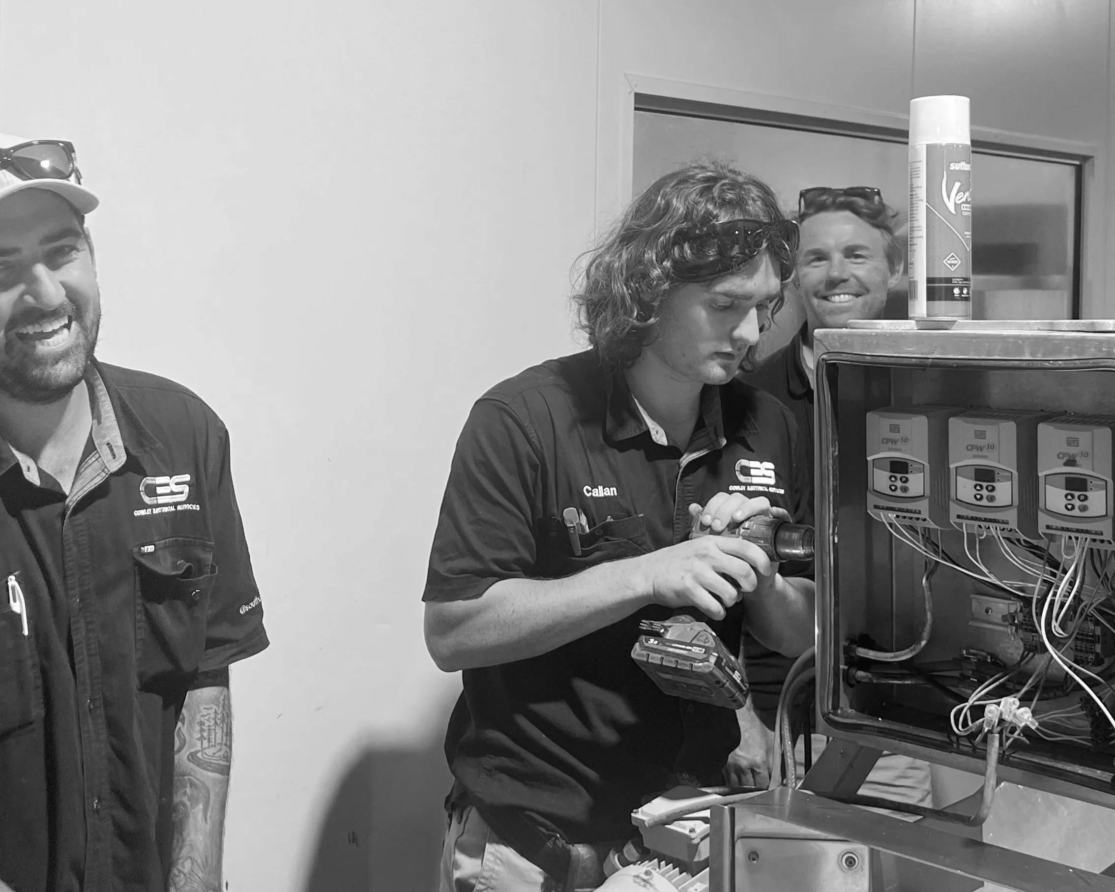 Four men in work uniforms with company logos, one with sunglasses on his hat, working on electrical equipment or control panels. One man is holding a tool, and others are observing or assisting. The room has plain walls and the equipment is mounted on a cart.
