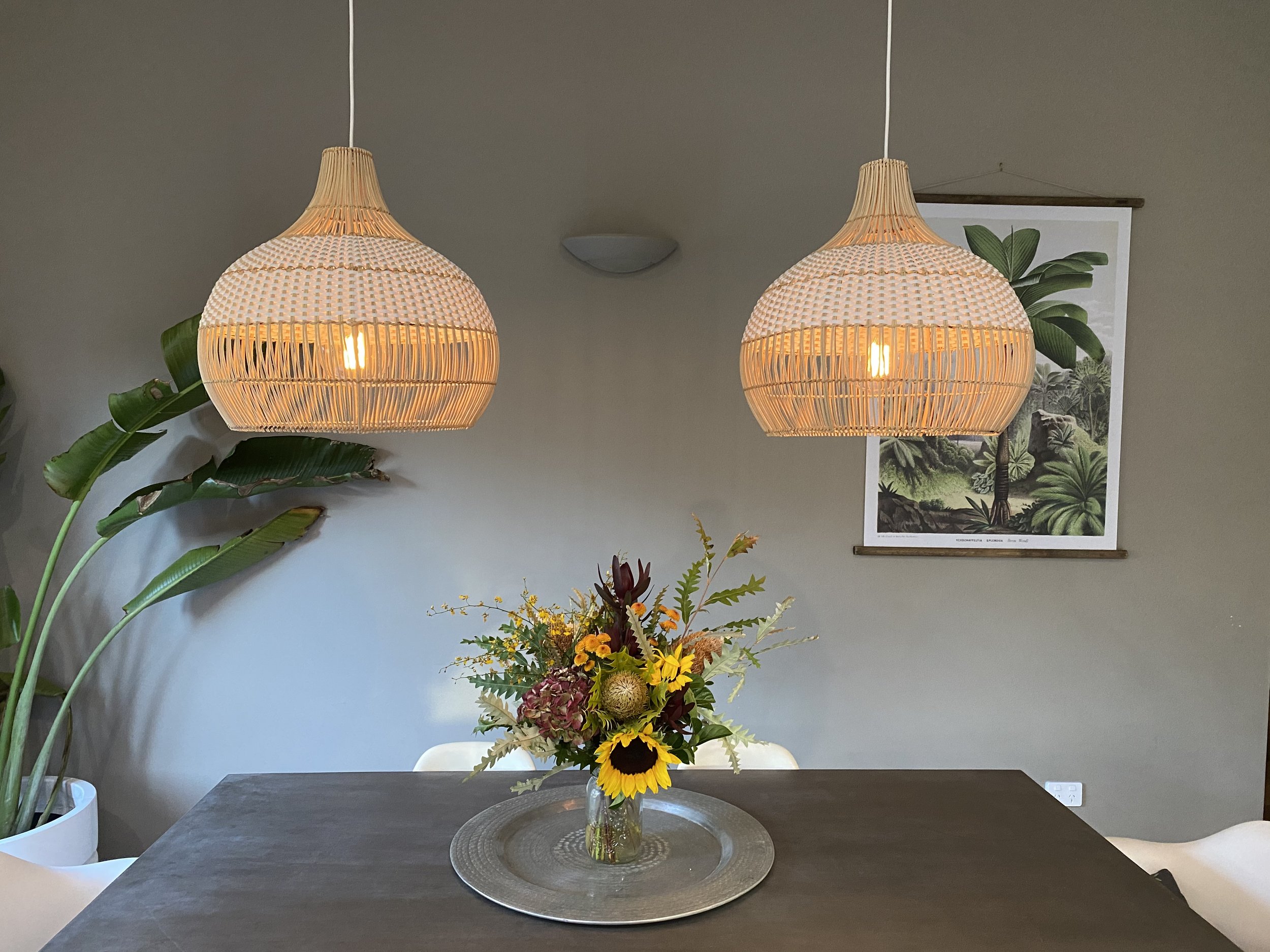 Two woven pendant lights hanging above a table with a floral arrangement of sunflowers and other flowers, with a plant and a tropical leaf print in the background.