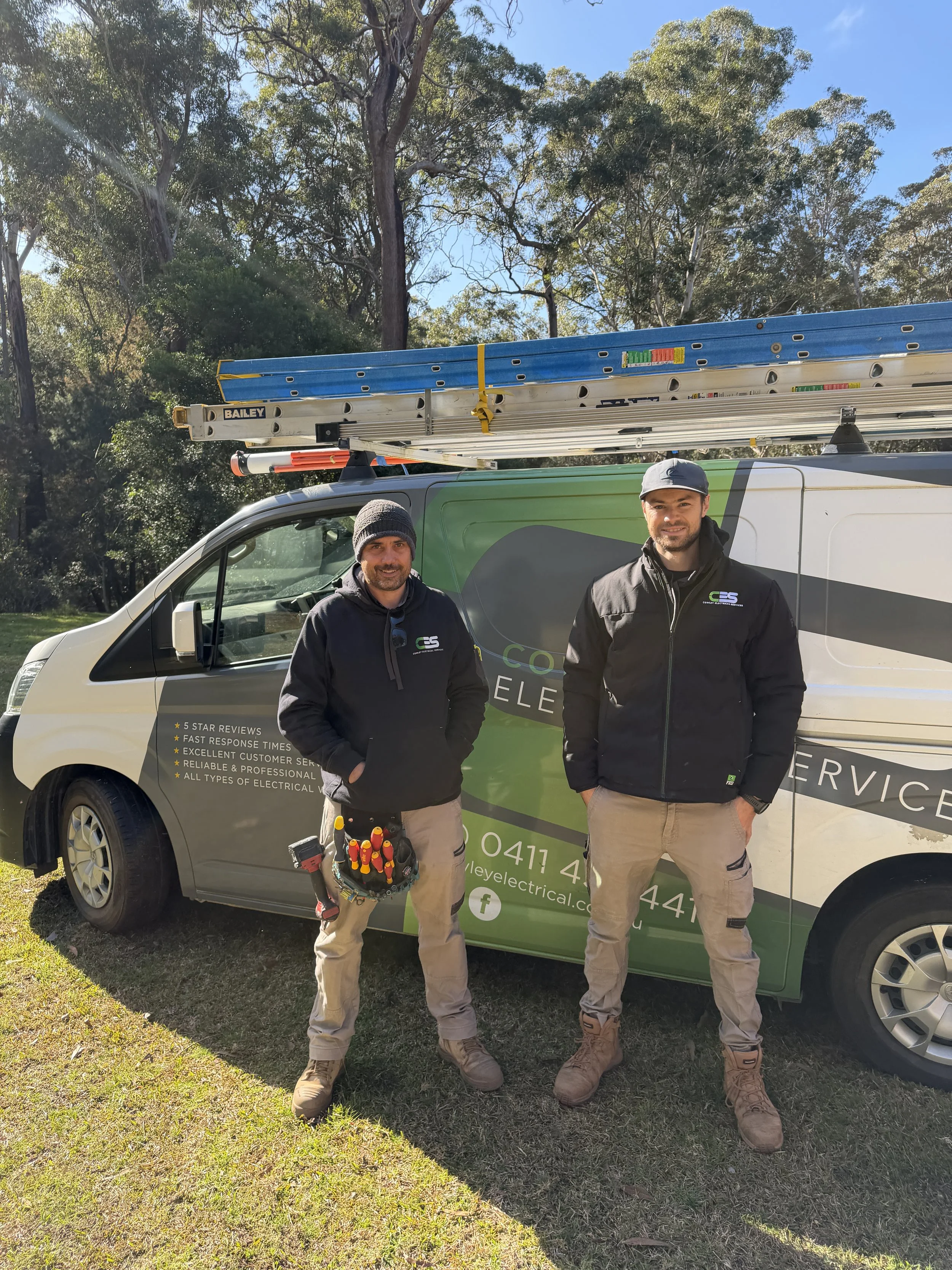 Two men standing next to a service van outdoors on grass, with trees and a blue sky in the background. One man has a tool belt and the other wears a cap. The van has signage related to electrical services.