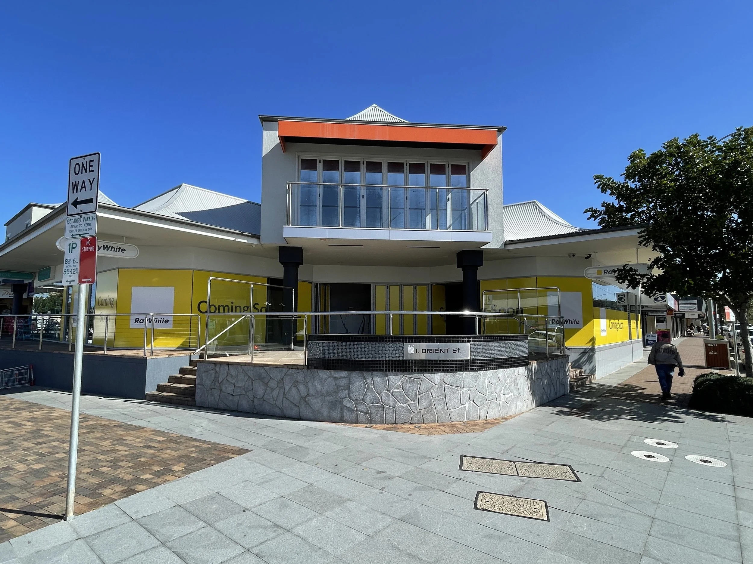A modern building with white walls, a small balcony, and a yellow sign that says "Coming Soon" on a sidewalk under a clear blue sky. A one-way street sign and some trees are visible in the scene.