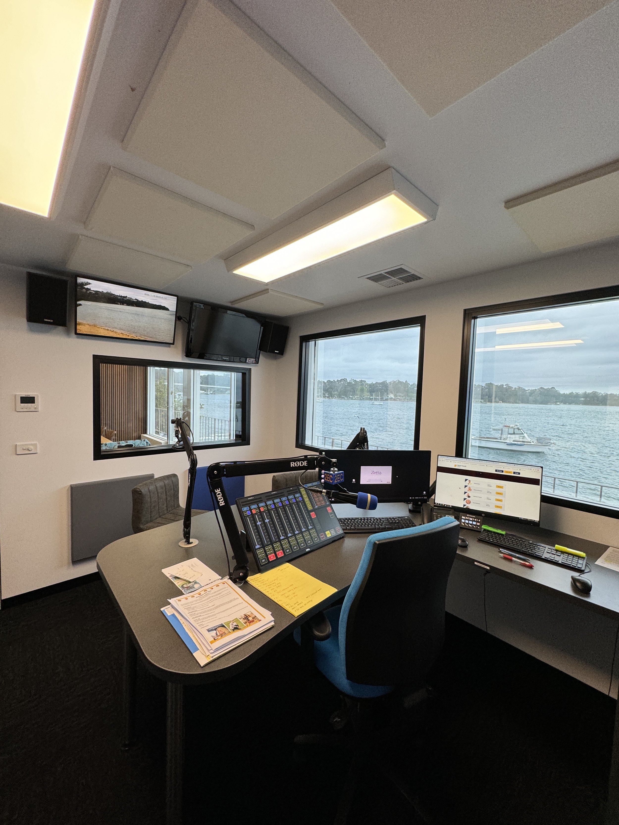 Radio studio with a large window overlooking a body of water, multiple computers and equipment on a desk, and a television mounted on the wall.