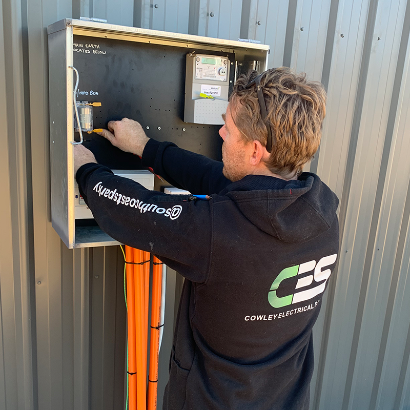 A man working on an electrical panel mounted on a metal wall, with orange conduit pipes hanging below.