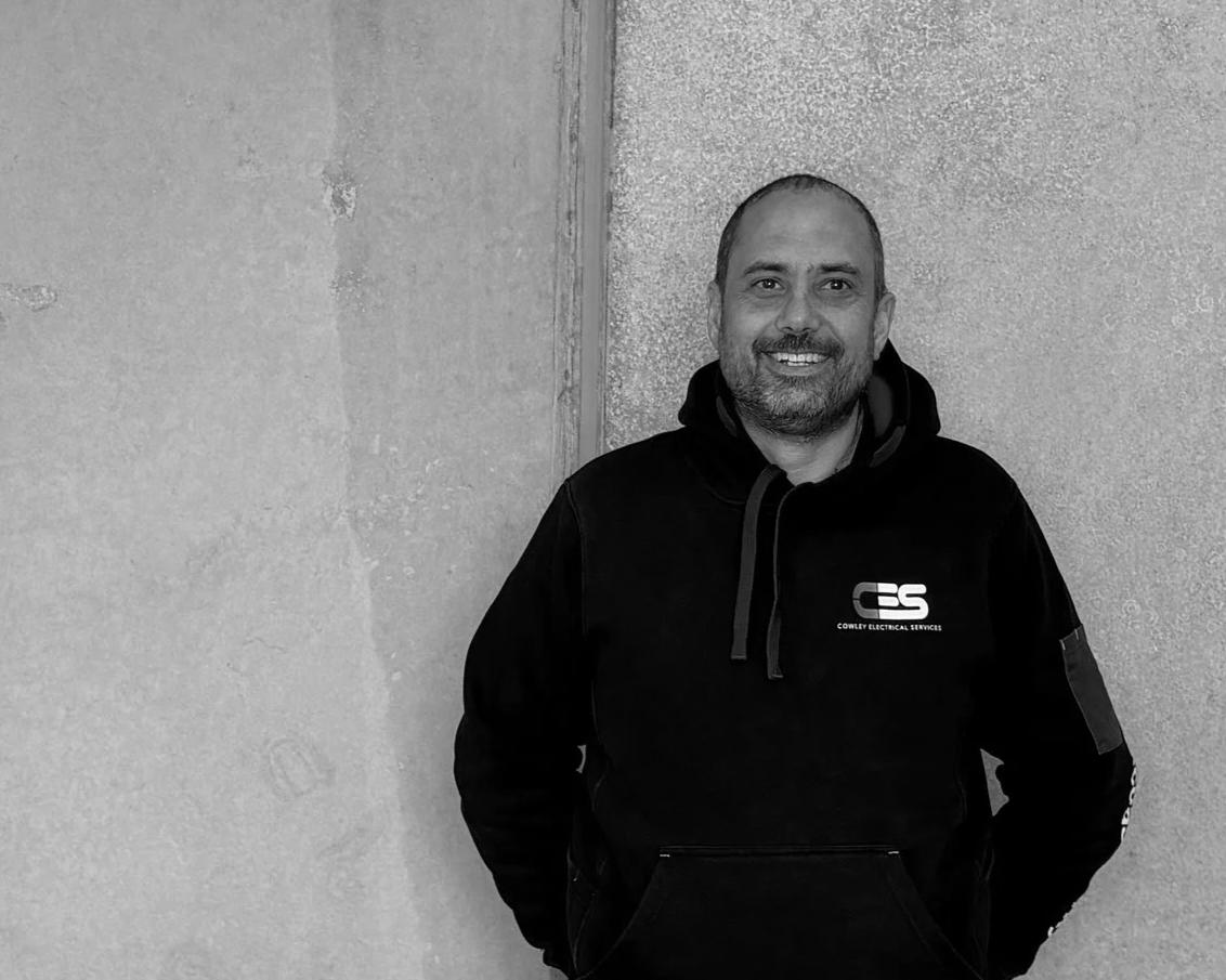 A smiling man with a beard and shaved head wearing a black hoodie with a logo and text that reads 'Cowley Electrical Services,' standing against a textured wall.