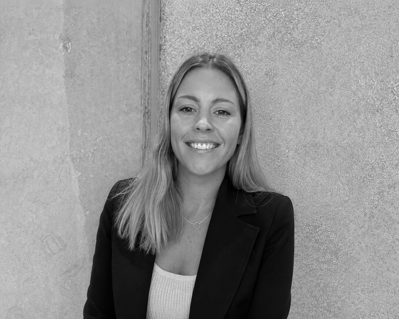 Black and white photo of a smiling woman with long hair, wearing a blazer, standing in front of textured wall.