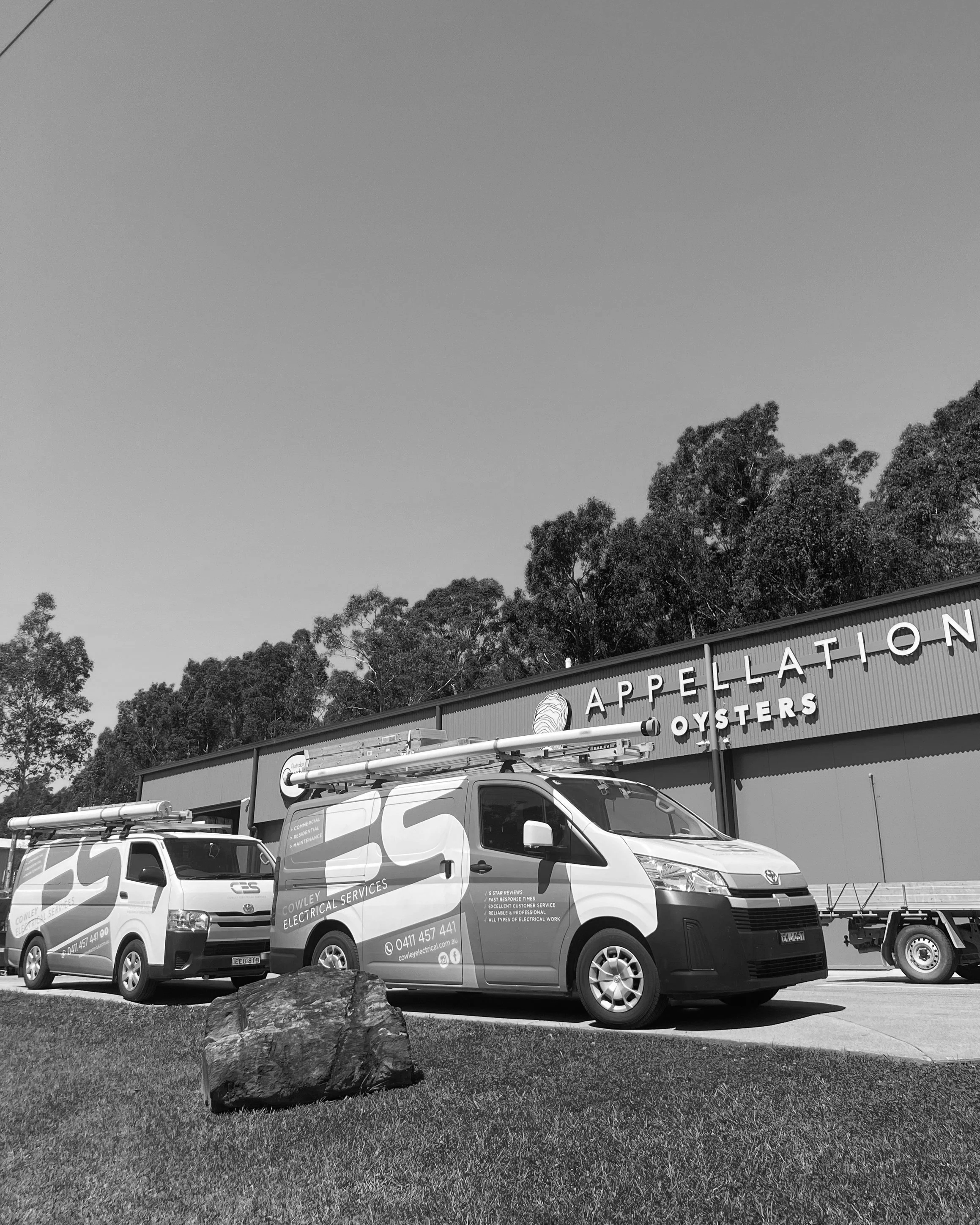 Two service vans parked in front of an electrical services business building. The vans have company branding and contact information, with a row of trees and part of the building's sign visible in the background.