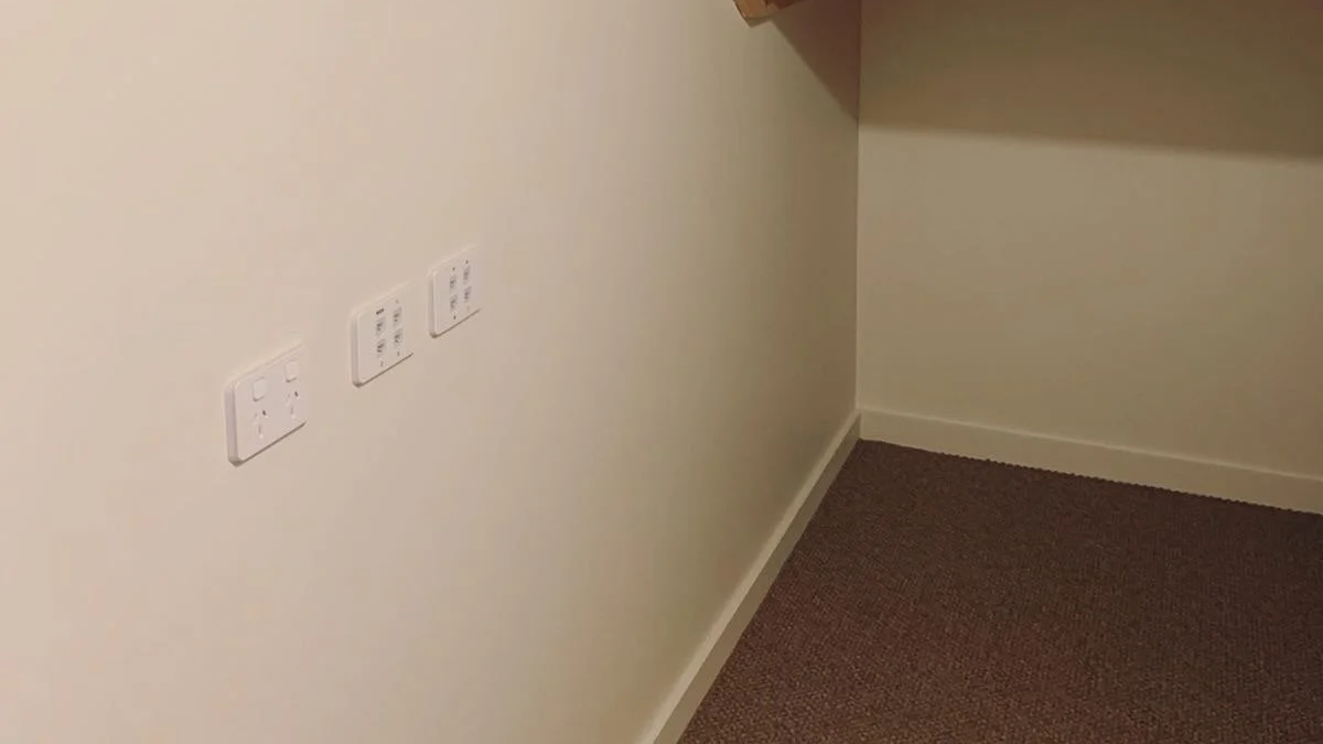 Empty corner of a room with three electrical outlets on a cream-colored wall and a brown carpeted floor.