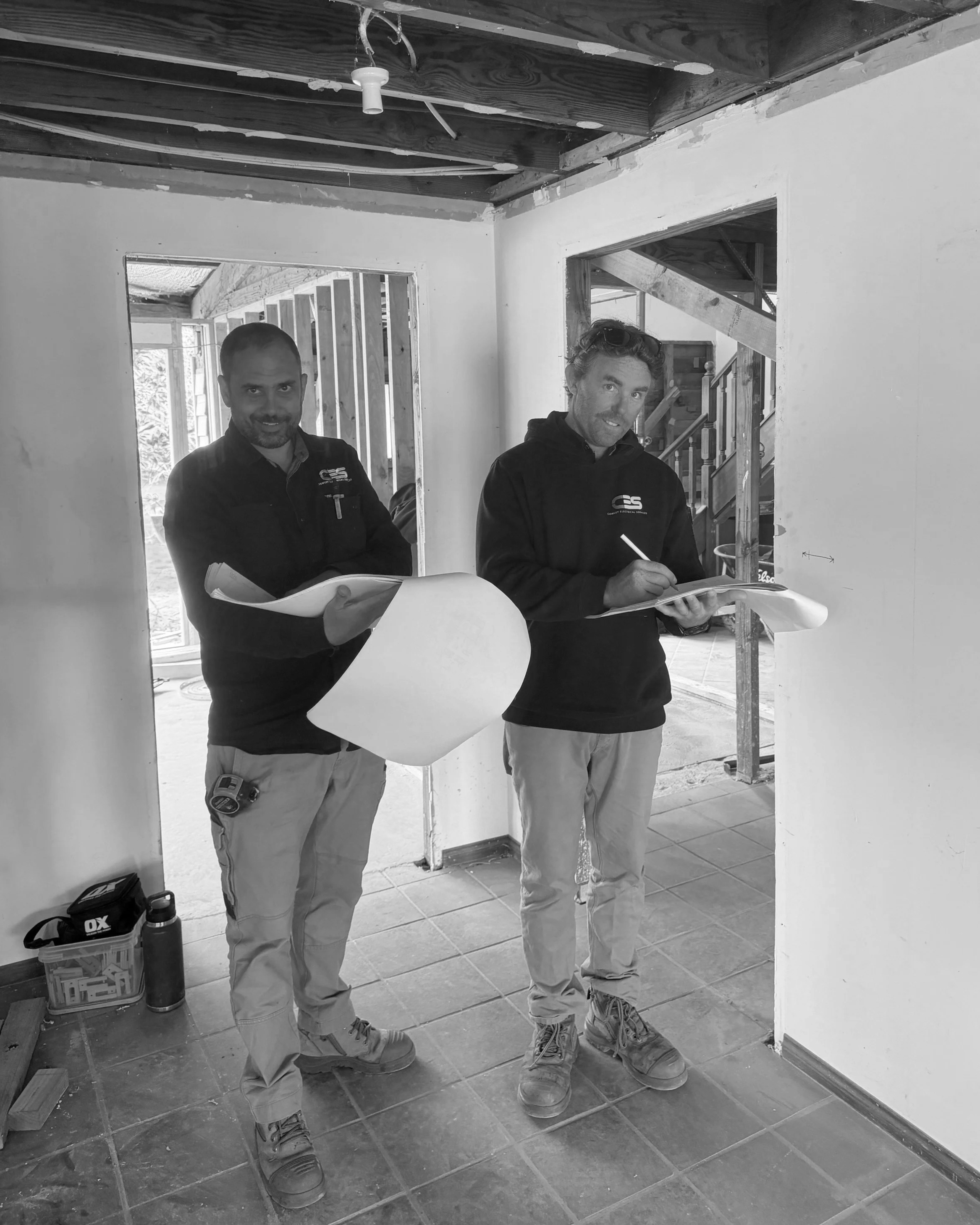 Two men in construction clothing standing inside a building under construction, holding blueprints, with building materials and tools on the floor, and open window frames behind them.