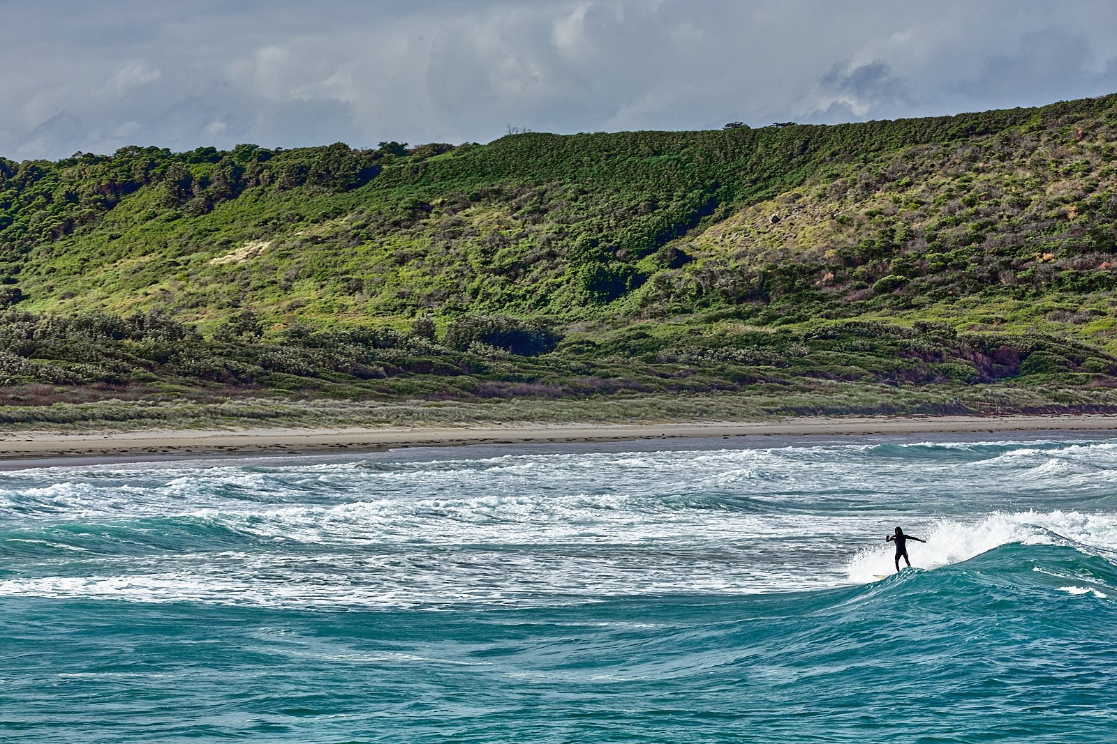 A person surfing on a wave in the ocean near a sandy beach and green hills in the background.