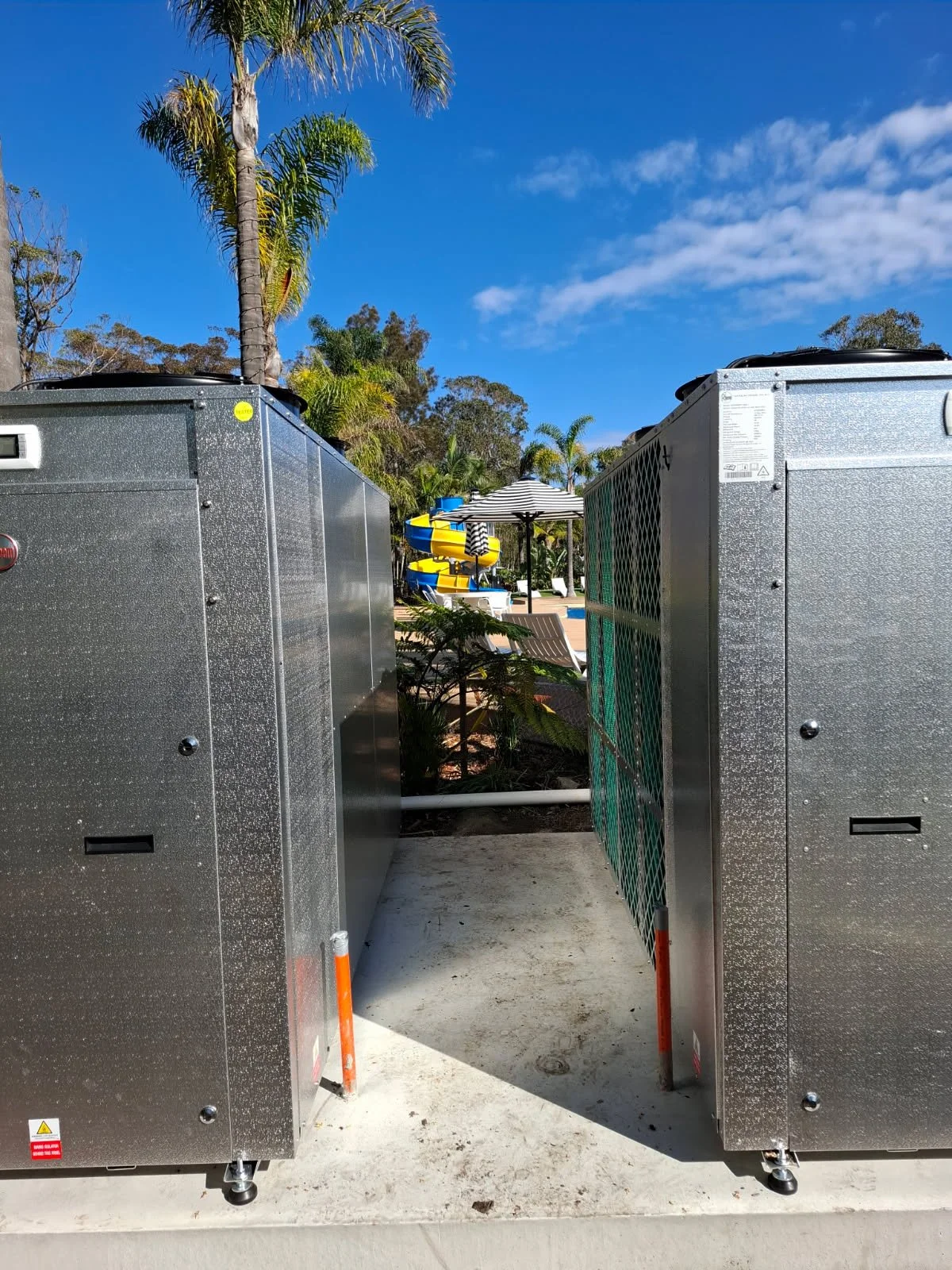 Two large metal air conditioning units with a pathway between them lead to a swimming pool area with lounge chairs, umbrellas, and water slides. Tall palm trees and a blue sky are in the background.