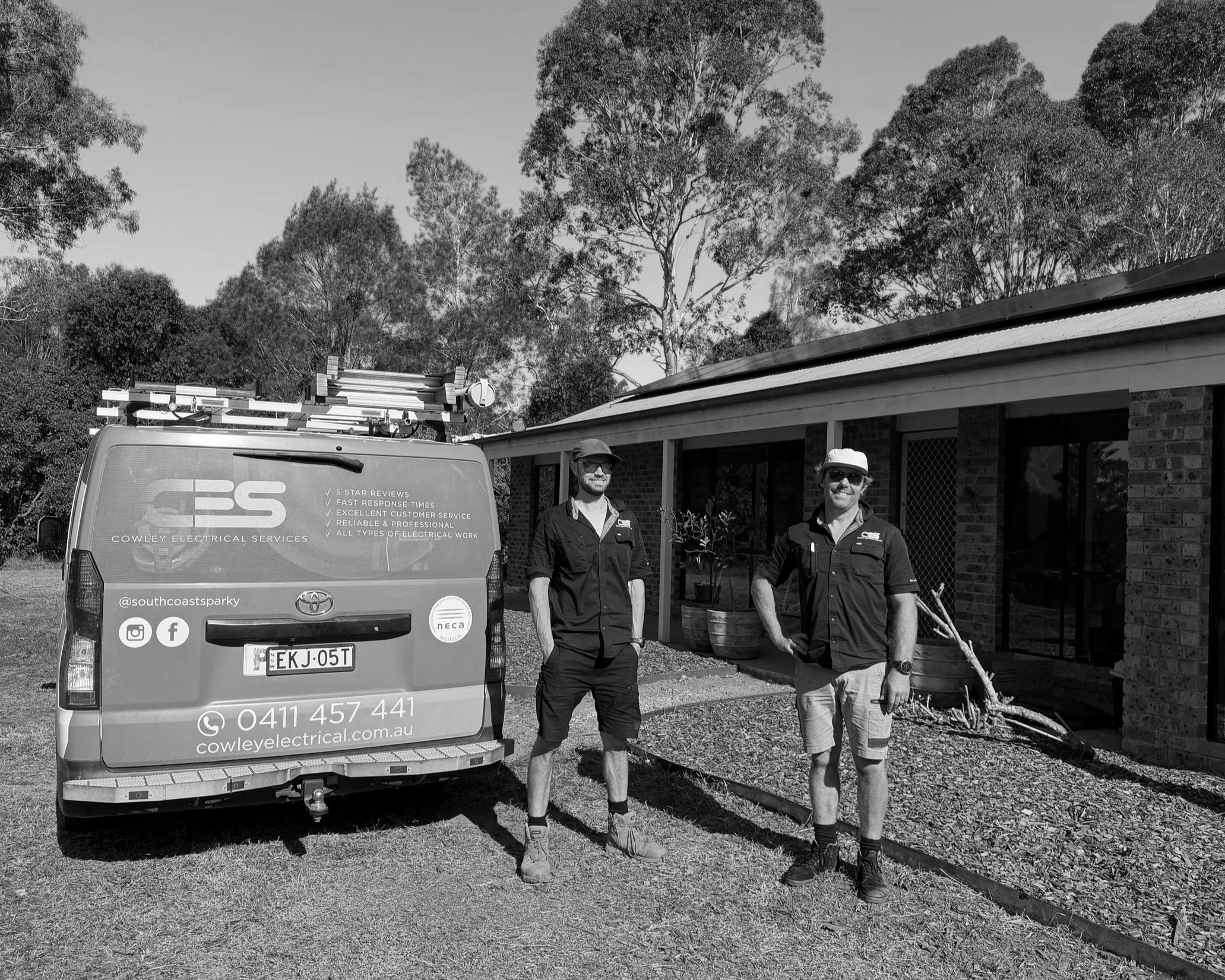 Three men standing outdoors beside a white van with business decals, in front of a house with a brick wall and a roof, surrounded by trees and shrubs, with a clear sky in the background. All three men are wearing dark work uniforms, with two wearing hats.