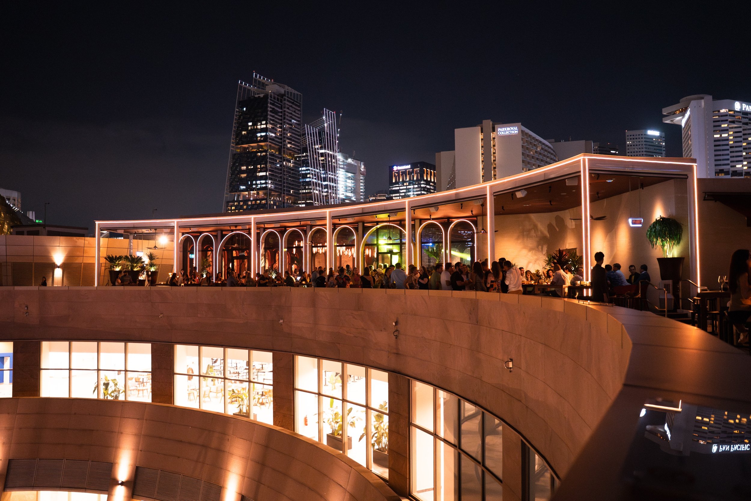 Night view of a rooftop restaurant in a modern city with skyscrapers, illuminated with string lights and filled with people dining, overlooking a city skyline.