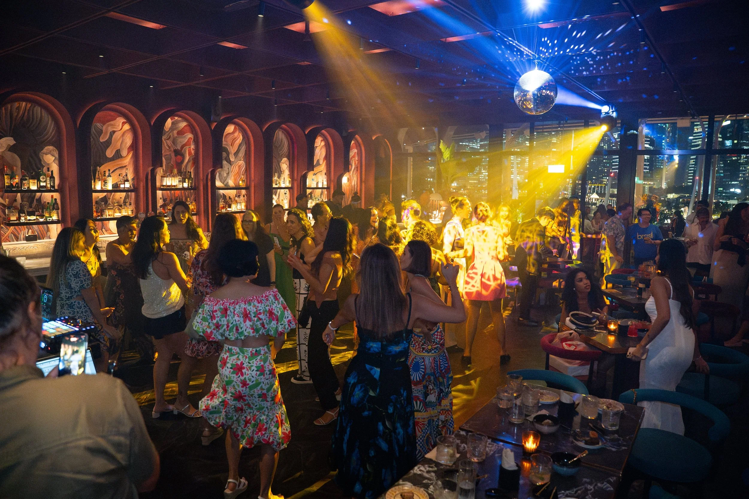People dancing and socializing in a lively nightclub with colorful lighting, a disco ball, and a city skyline visible through large windows.