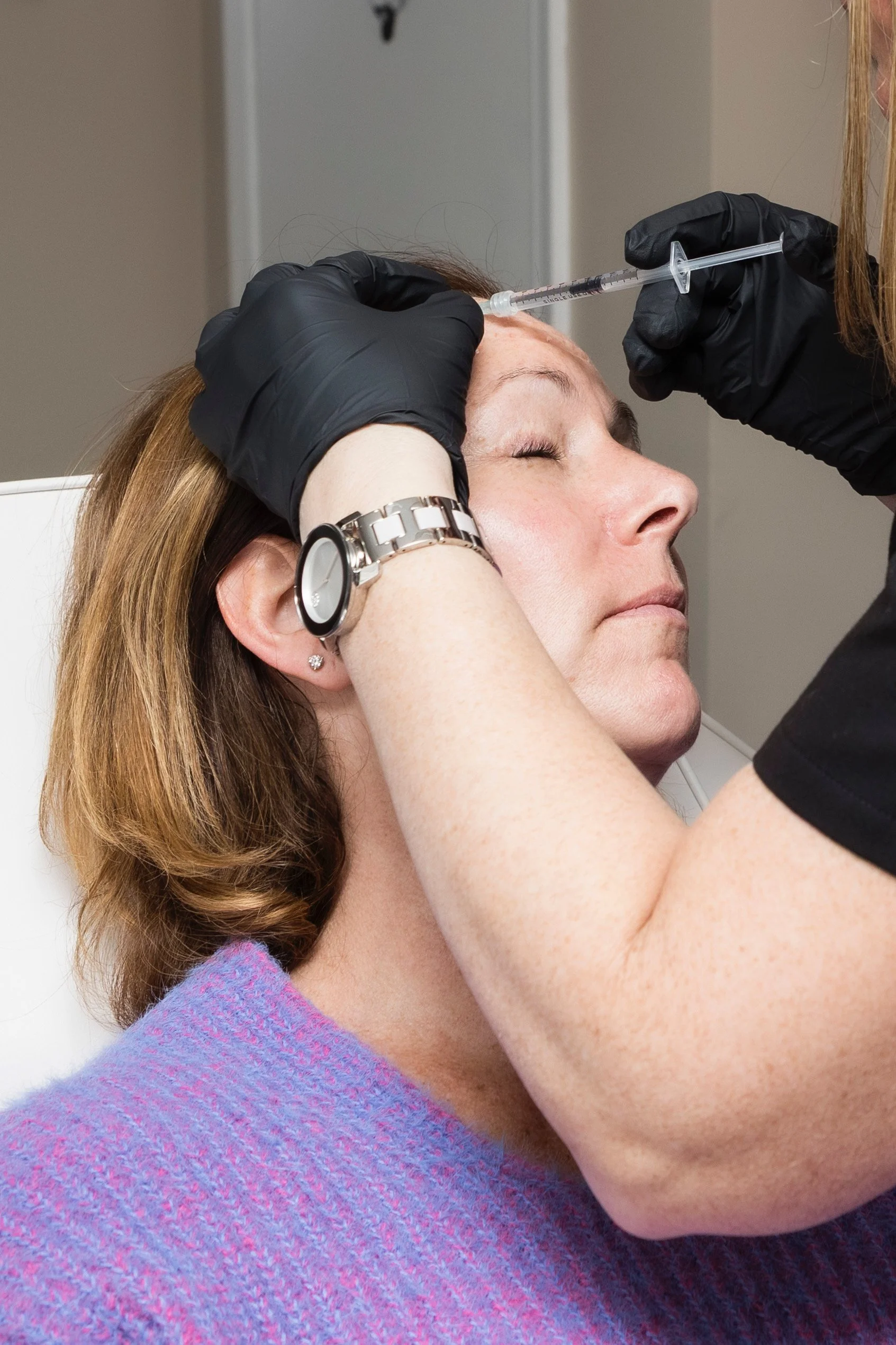 A healthcare professional administers a facial injection to a woman with closed eyes, using a syringe while wearing black gloves.