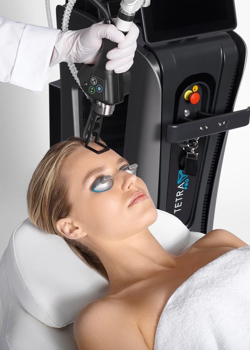 A woman receiving laser skin treatment on her forehead while lying on a treatment bed with protective eye shields in a medical clinic.